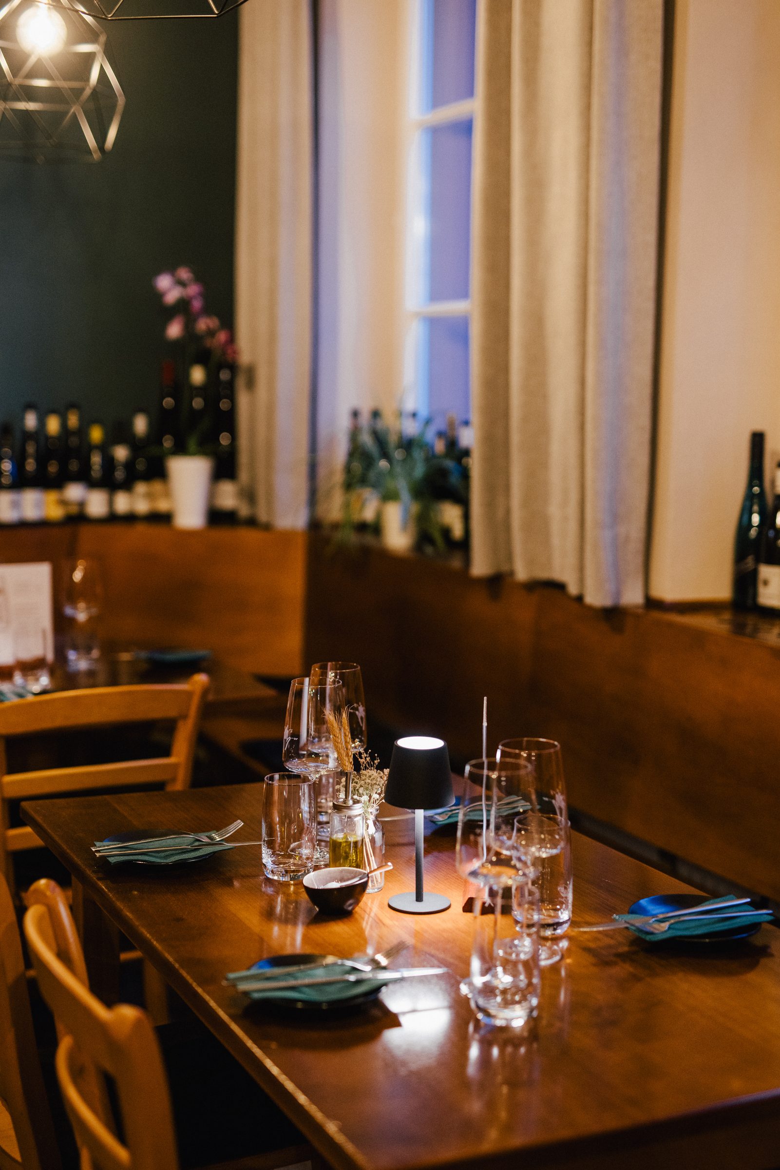 Gedeckter Tisch mit Weingläsern und Tischleuchte am Fenster im Restaurant 5 Bäuerlein, dahinter Weinflaschen und Orchideen