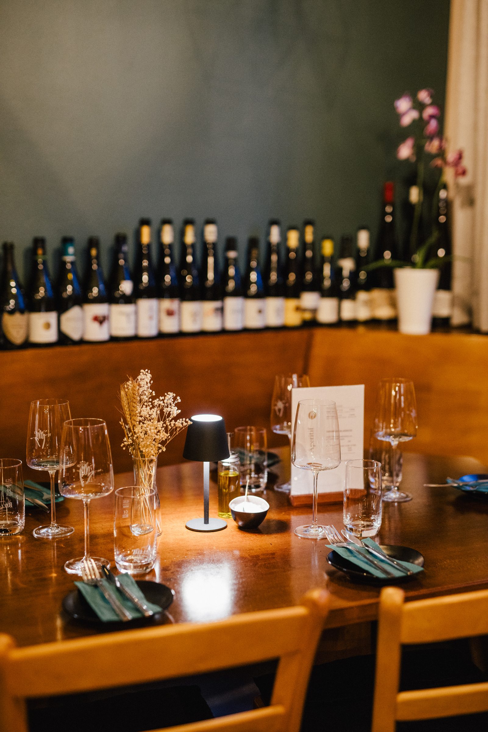 Gedeckter Ecktisch mit Tischleuchte, Trockenblumen und Menükarte vor einem Weinregal an dunkelgrüner Wand im Restaurant 5 Bäuerlein