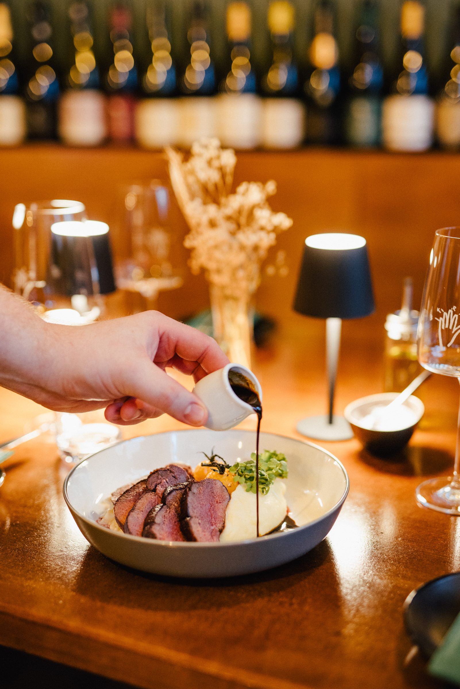 Hand gießt dunkle Sauce über rosa gebratenes Fleisch in einer Schale, dahinter Tischleuchten und ein Weinregal im Restaurant 5 Bäuerlein
