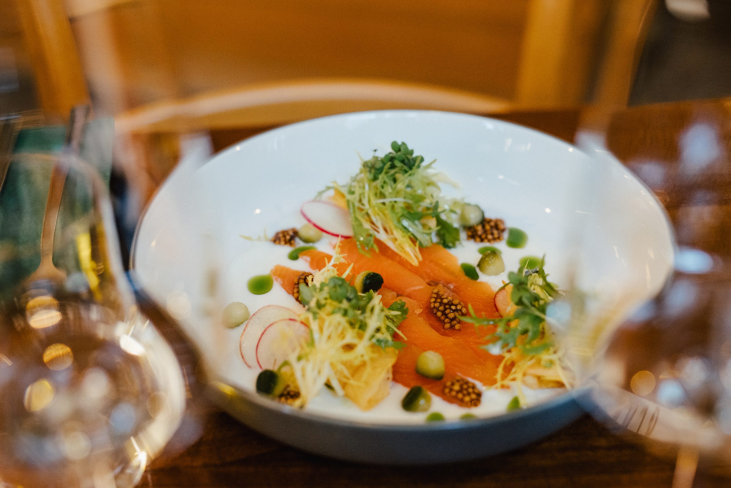 Vorspeise mit Lachs, Radieschen, Kresse und Senfkörnern auf einem weißen Teller, im Vordergrund unscharf Weingläser