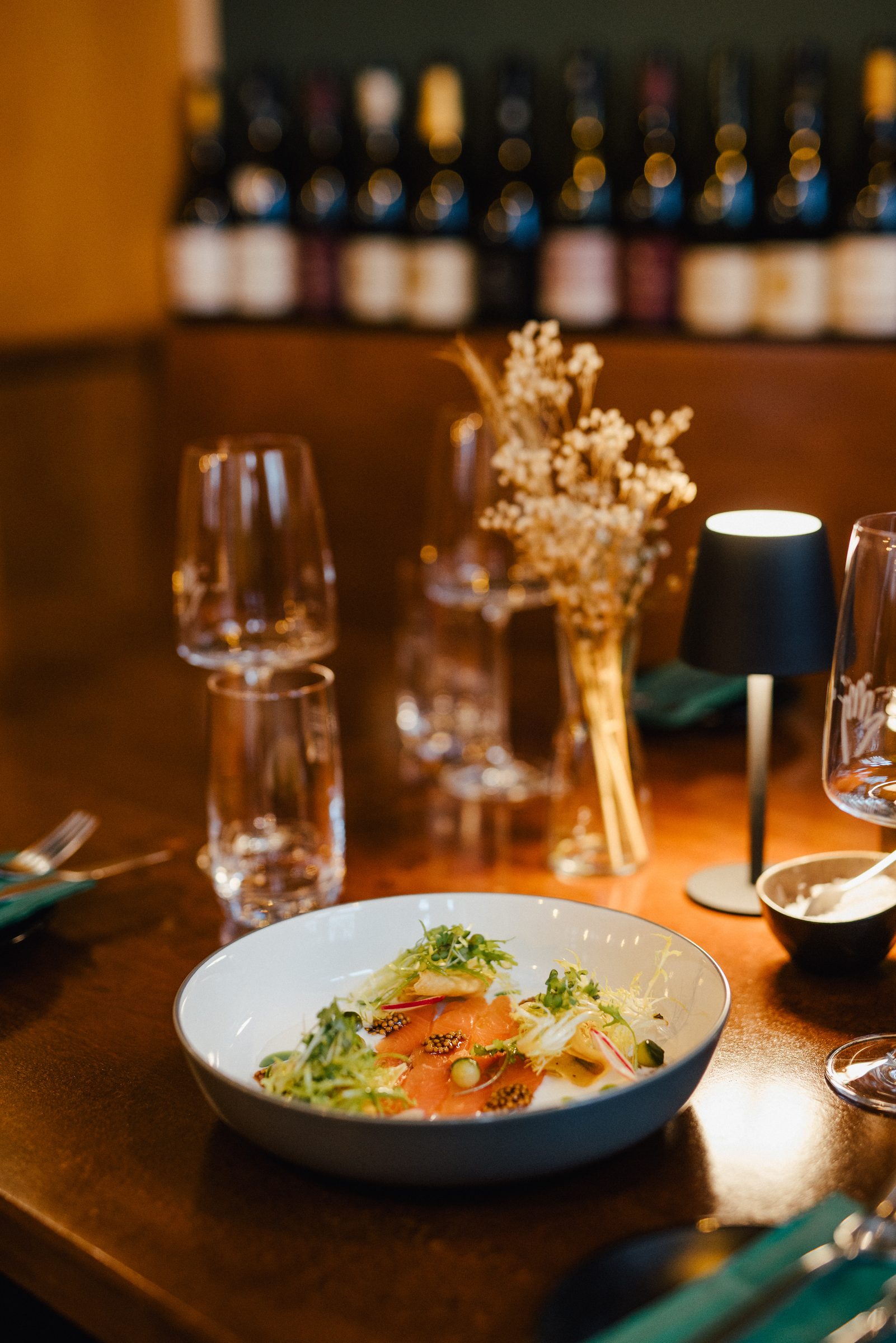 Vorspeise mit Lachs in einer weißen Schale auf einem gedeckten Tisch, dahinter Tischleuchte, Trockenblumen und ein Weinregal im Restaurant 5 Bäuerlein
