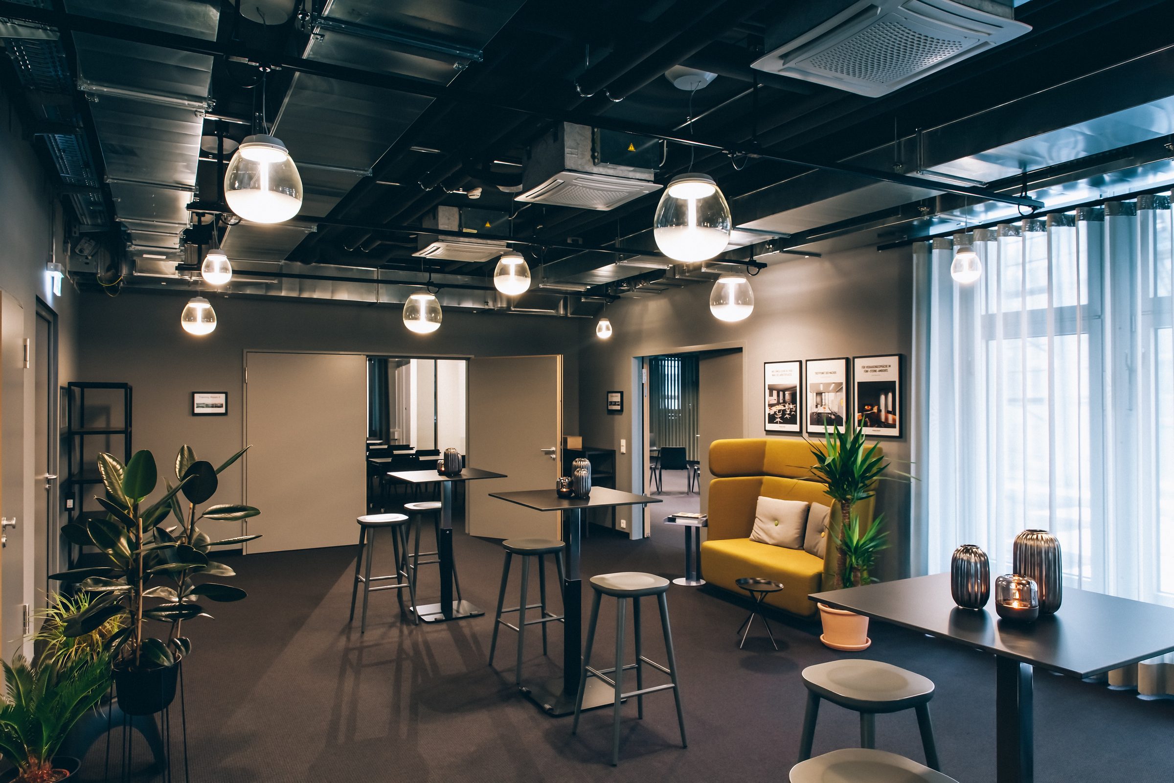 Lounge der Design Offices Stuttgart mit Pflanzen, Stehtischen, gelbem Sofa und Pendelleuchten von Artemide