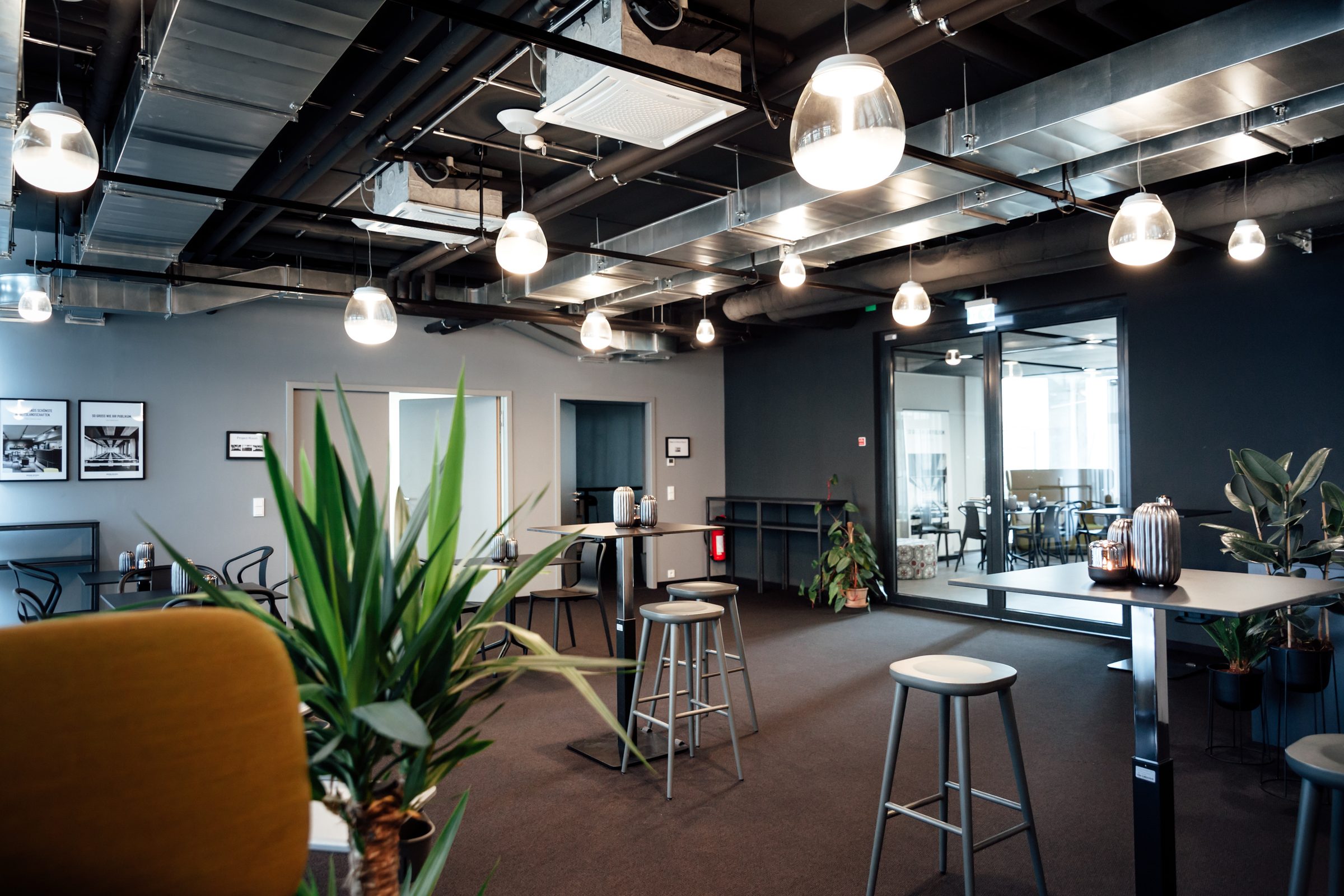 Pendelleuchten von Artemide über Stehtischen in der Lounge der Design Offices Stuttgart, im Vordergrund eine Pflanze und ein gelber Sessel