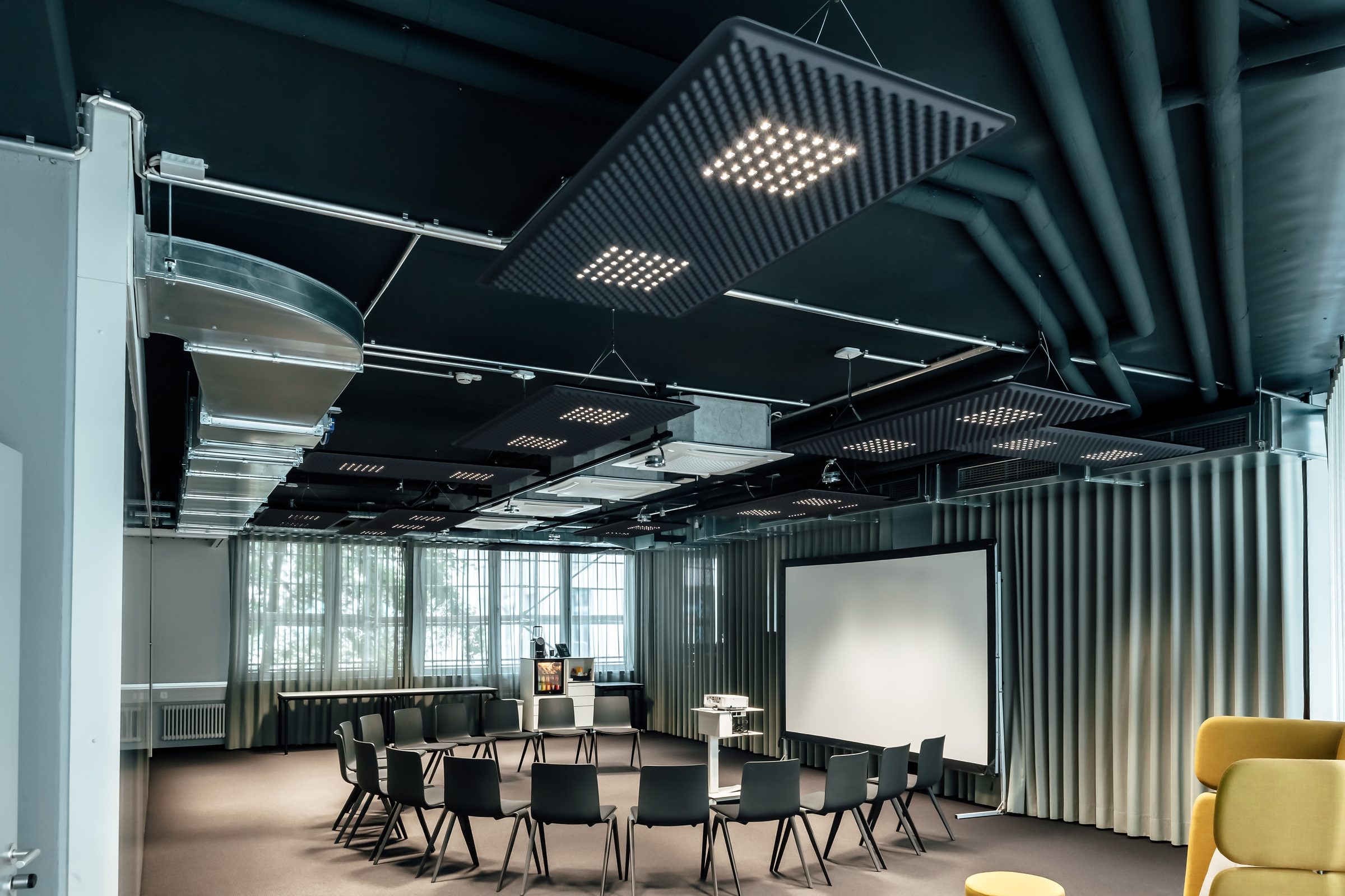 Konferenzraum mit Stuhlkreis und Leinwand unter Akustikpaneelen mit LED-Leuchten, im Vordergrund ein gelber Sessel, Design Offices Stuttgart