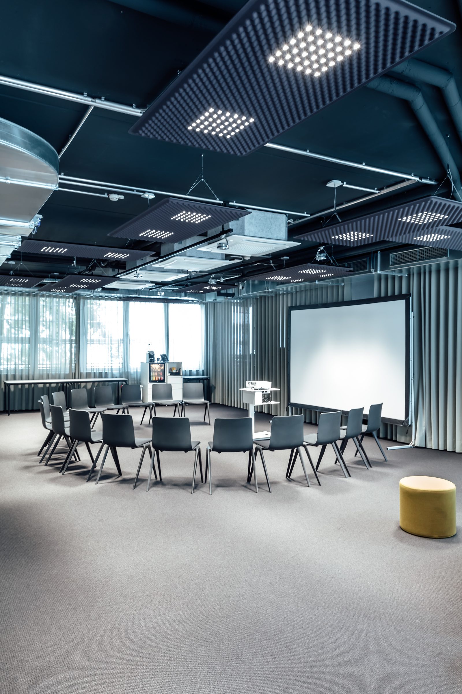 Konferenzraum mit Stuhlkreis, Leinwand und gelbem Hocker unter Deckenpaneelen mit LED-Leuchten, Design Offices Stuttgart