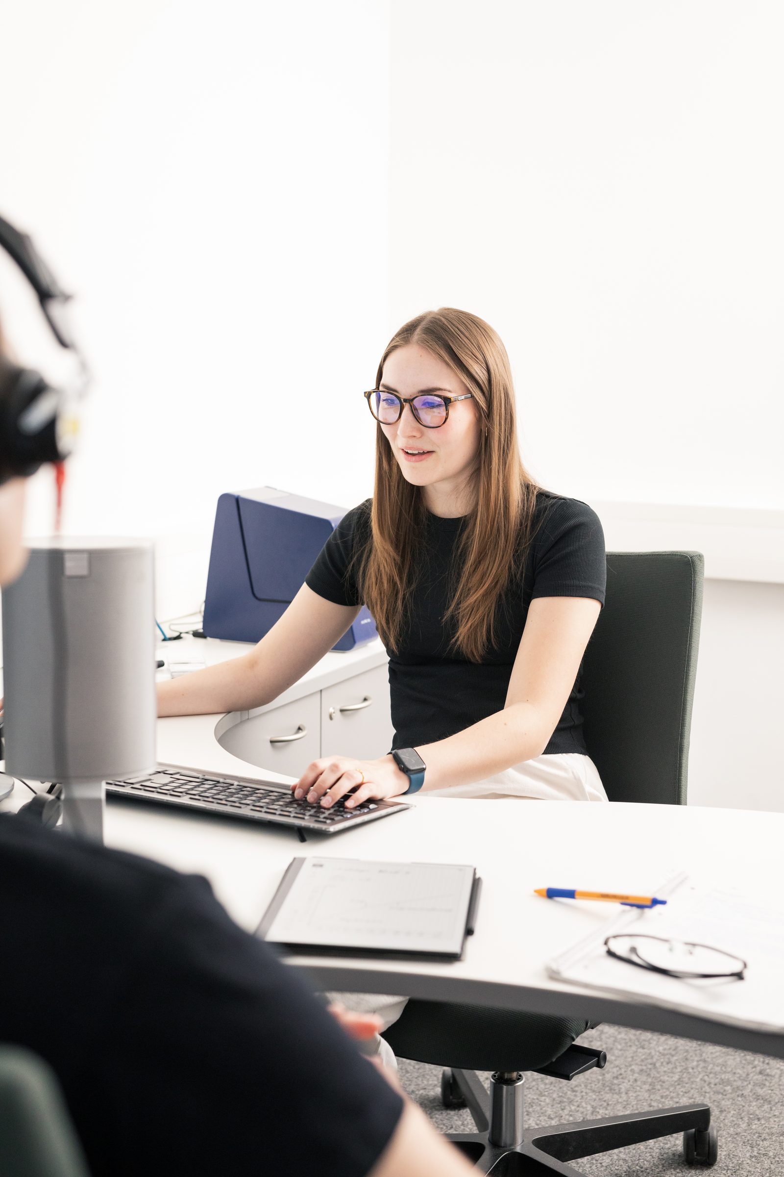 Junge Frau am Computer bei einer Hörtest-Übung, im Vordergrund eine Person mit Kopfhörern