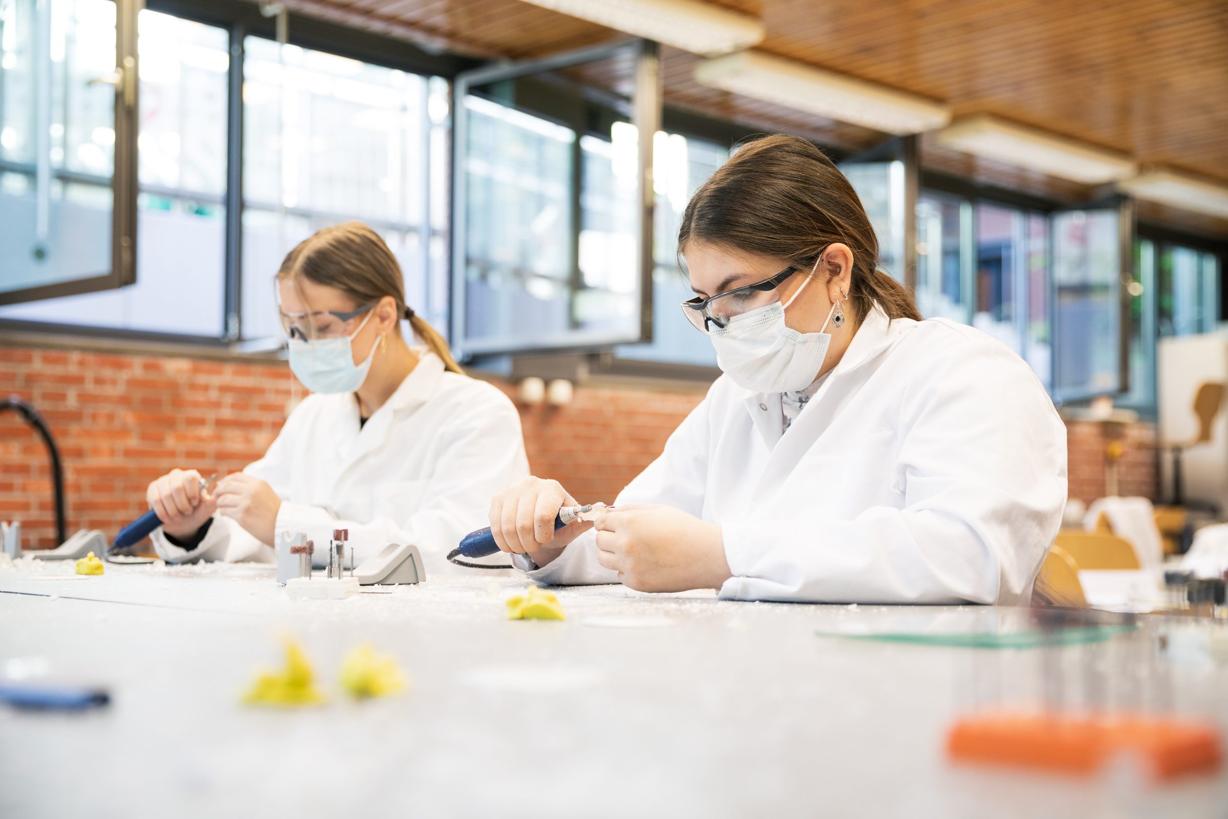Zwei Auszubildende in weißen Kitteln mit Schutzbrille und Maske arbeiten mit Schleifgeräten an einem Werktisch bei BIHA Hörakustik