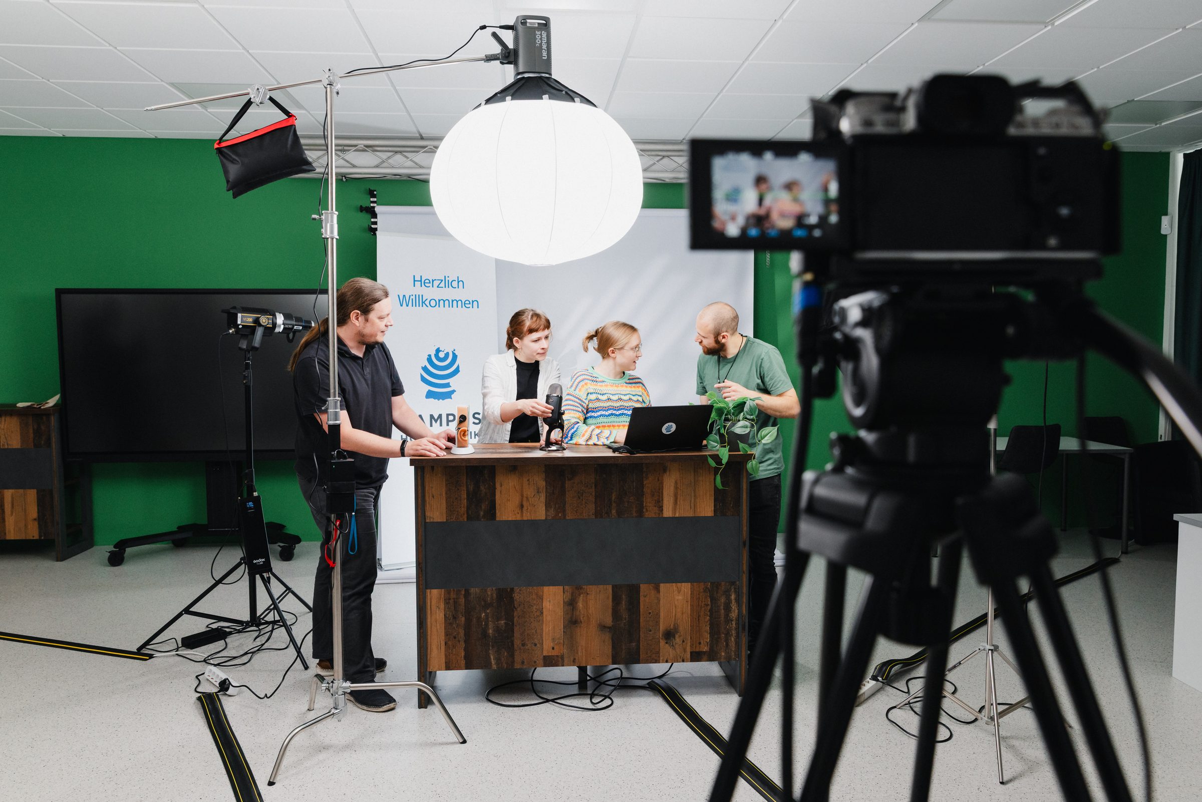 Videodreh im Studio von BIHA Hörakustik, vier Personen an einem Holztresen vor grüner Wand, vorn eine Kamera auf einem Stativ
