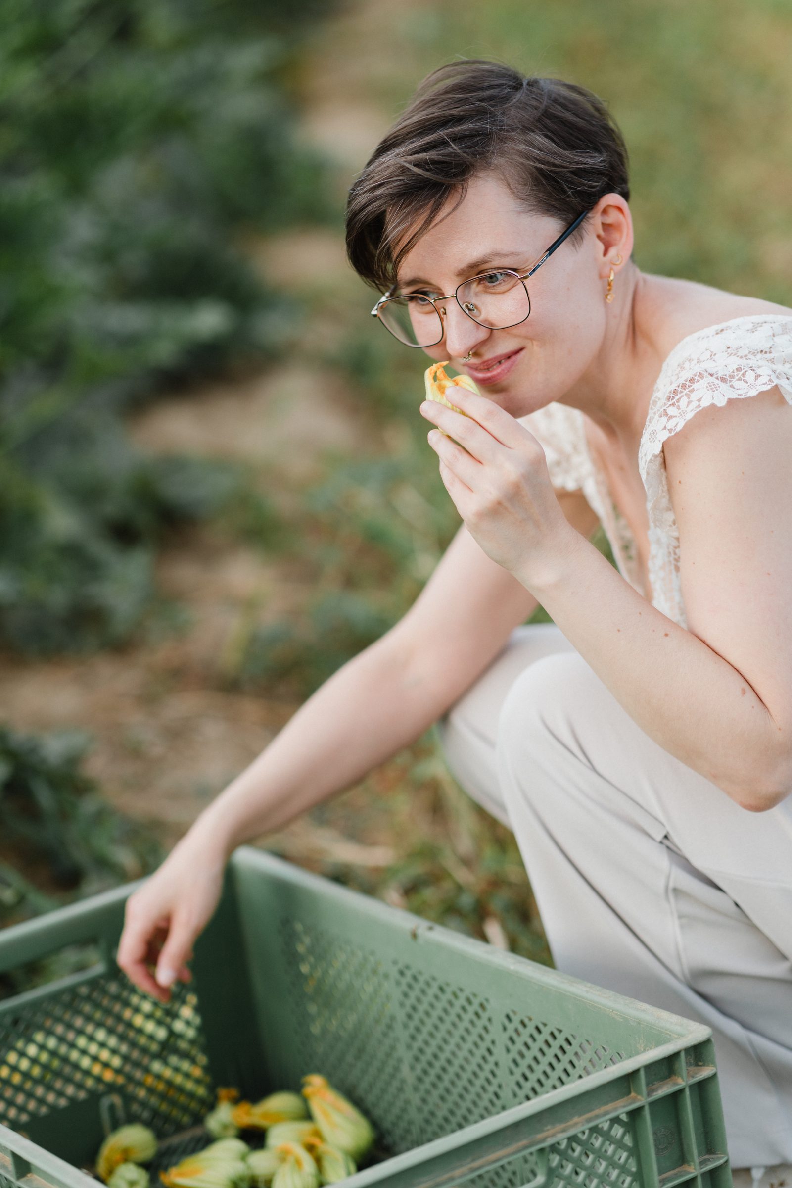 Frau mit Brille riecht an einer gelben Blüte, vor ihr eine grüne Erntekiste, Team von Colli Natur Friseure