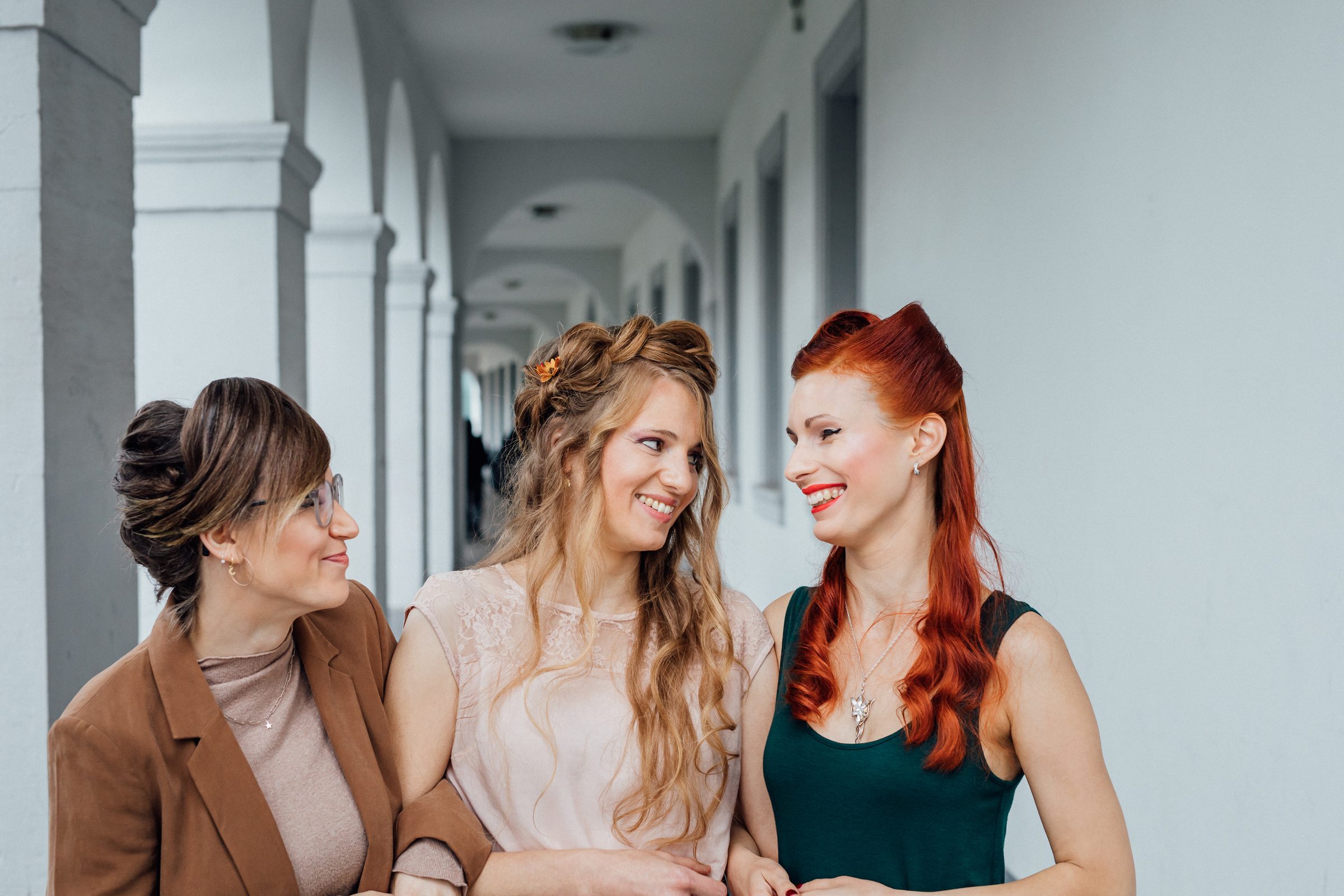 Drei Frauen mit aufwendig gestylten Frisuren lachen gemeinsam in einem hellen Säulengang, Fotoshooting für Colli Natur Friseure