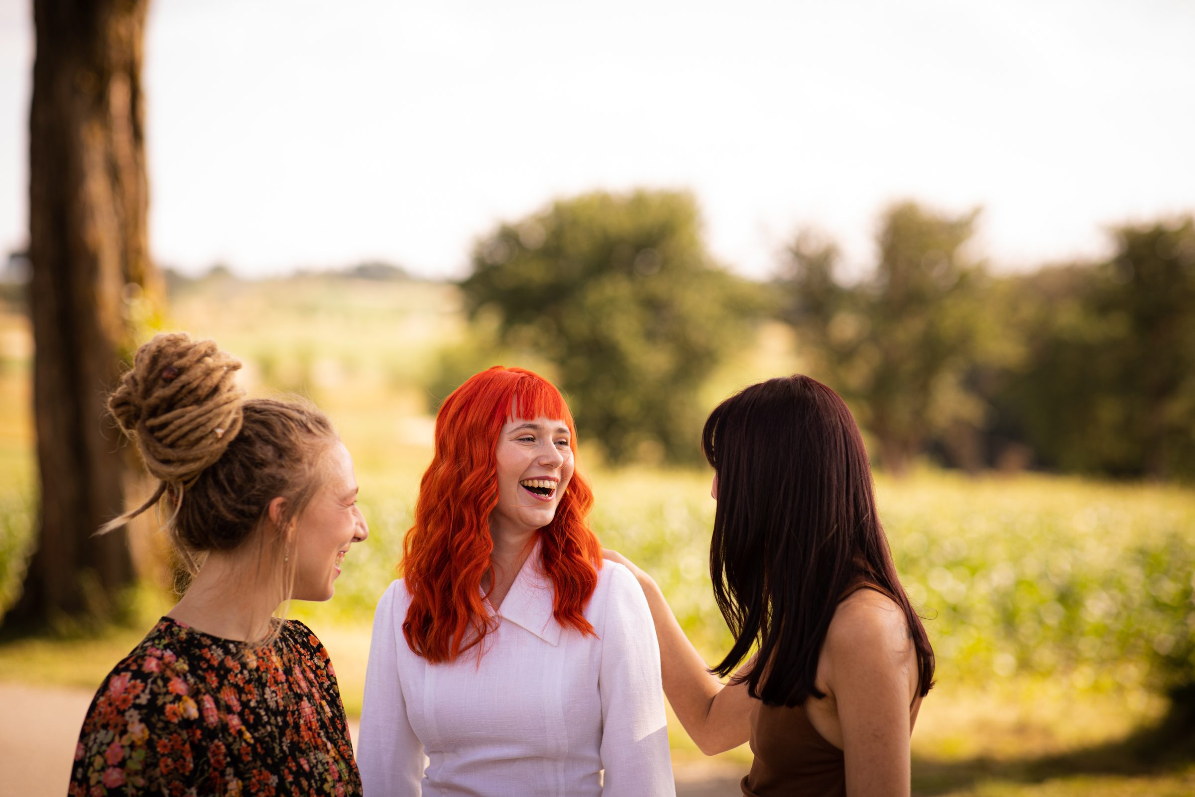 Drei Frauen mit Dreadlocks, roten Haaren und langen dunklen Haaren unterhalten sich lachend auf einem sonnigen Feld, Fotoshooting für Colli Natur Friseure