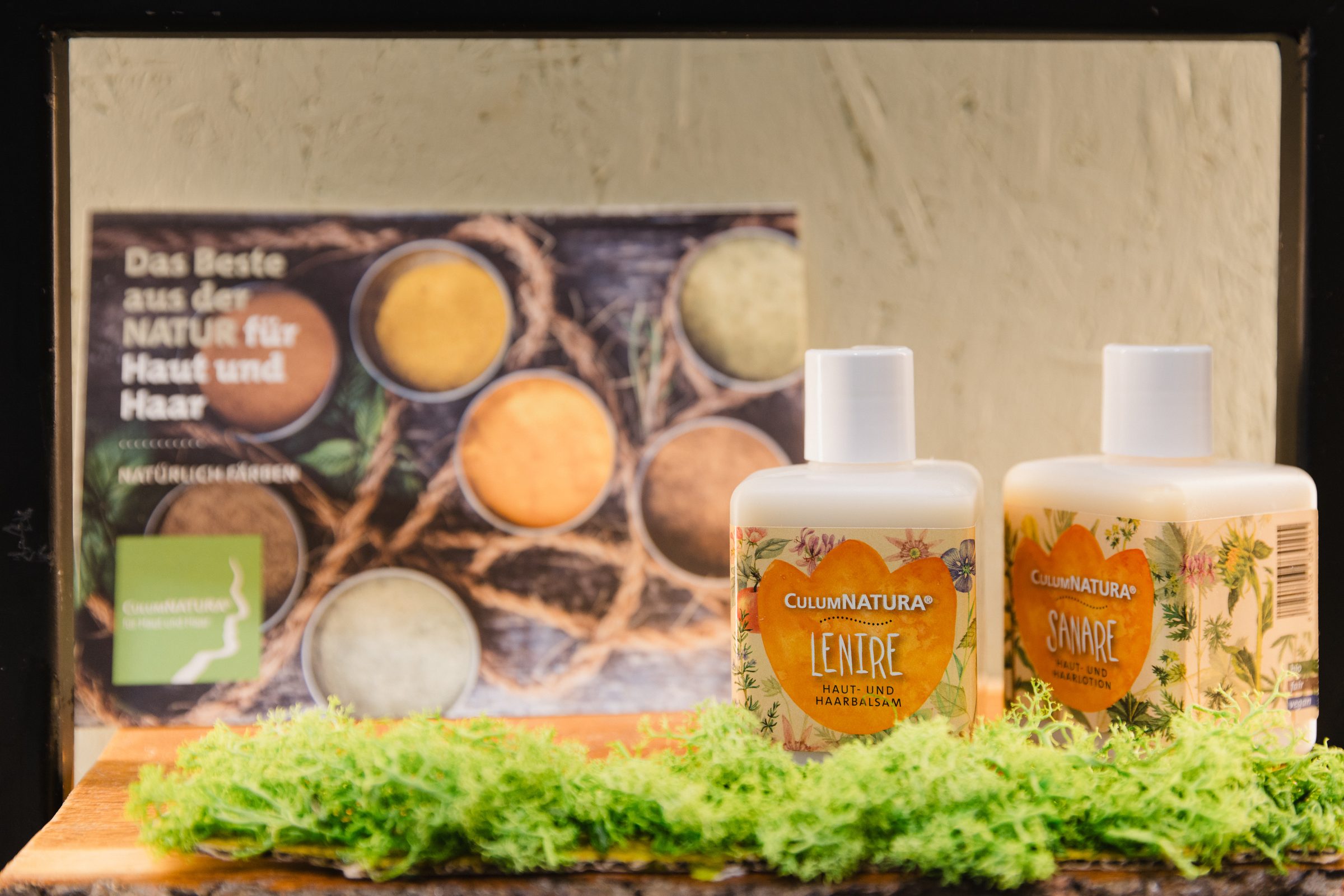 Zwei Flaschen Naturkosmetik für Haut und Haar auf grünem Moos, dahinter ein Plakat mit Schalen voller Pflanzenpulver, bei Colli Natur Friseure
