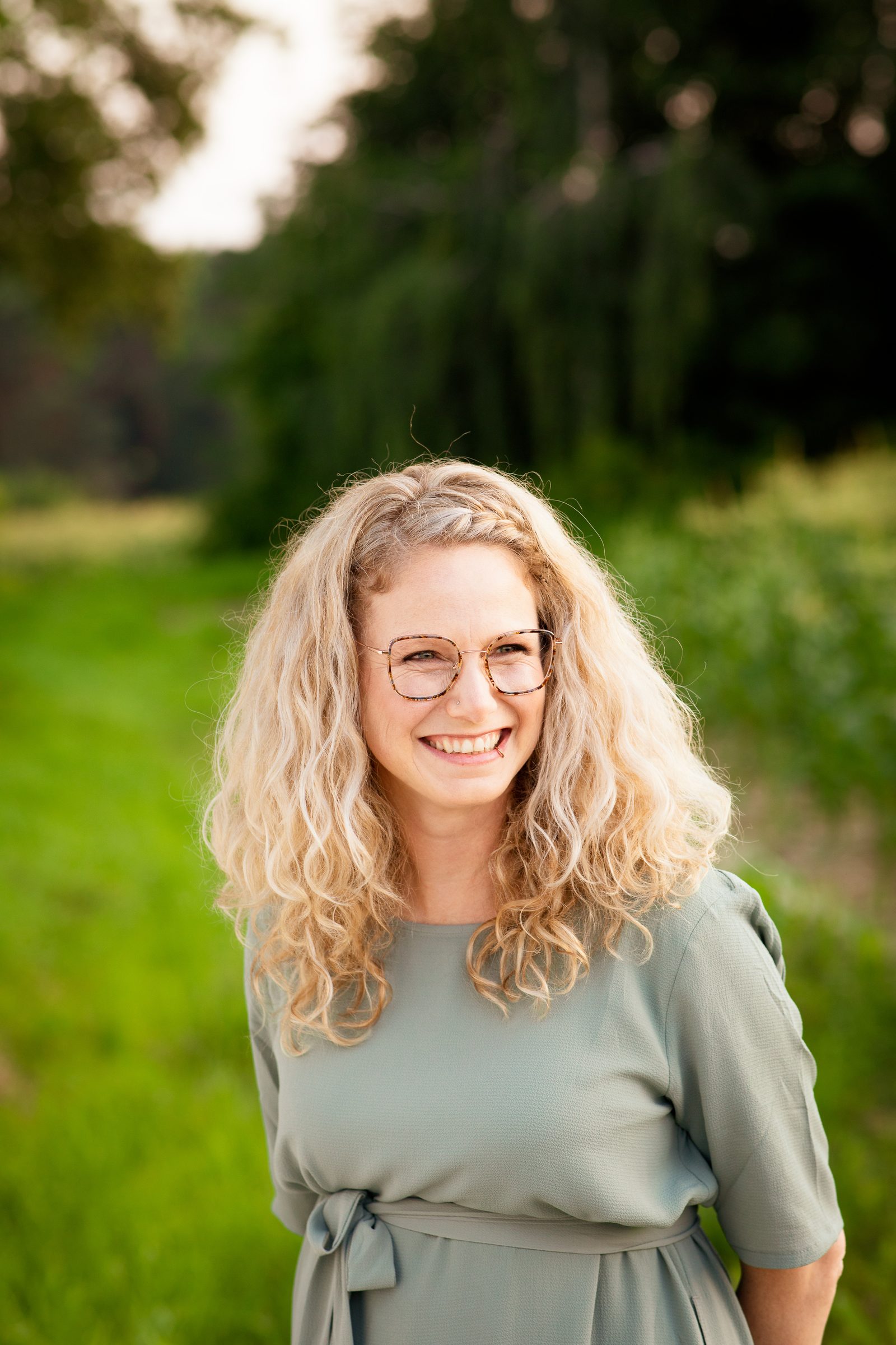 Frau mit langen blonden Locken und Brille lacht vor einer grünen Wiese, Fotoshooting für Colli Natur Friseure