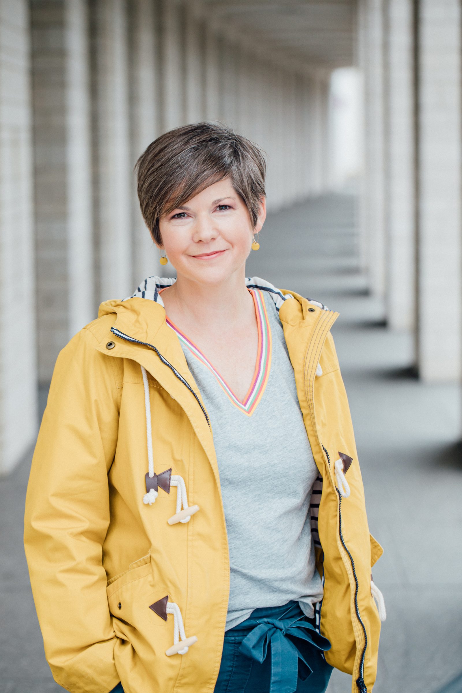 Frau mit kurzen braunen Haaren in gelber Regenjacke lächelt in einem hellen Säulengang, Fotoshooting für Colli Natur Friseure