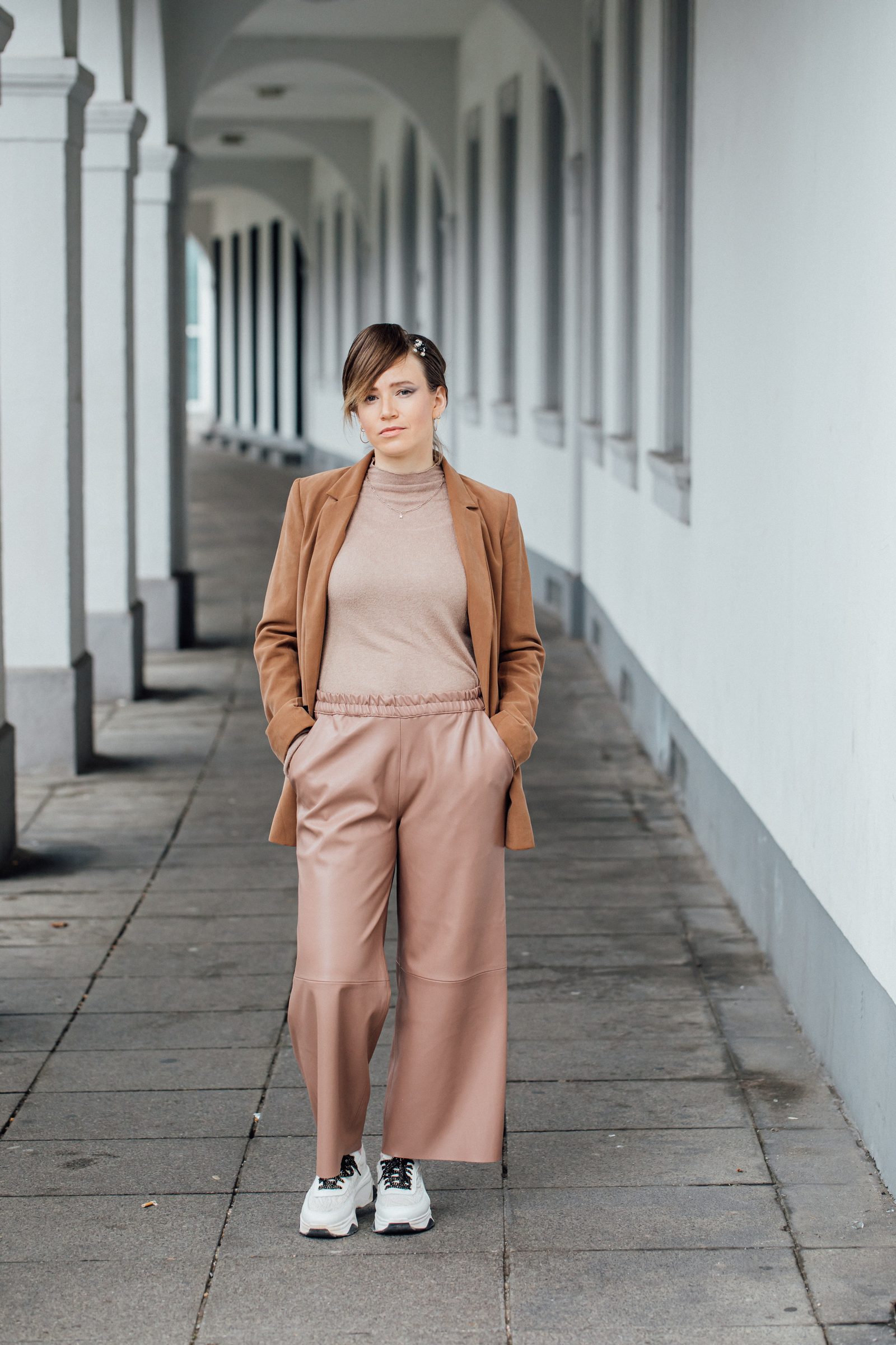 Frau in rosa Lederhose und brauner Jacke steht mit den Händen in den Taschen in einem langen Säulengang, Fotoshooting für Colli Natur Friseure
