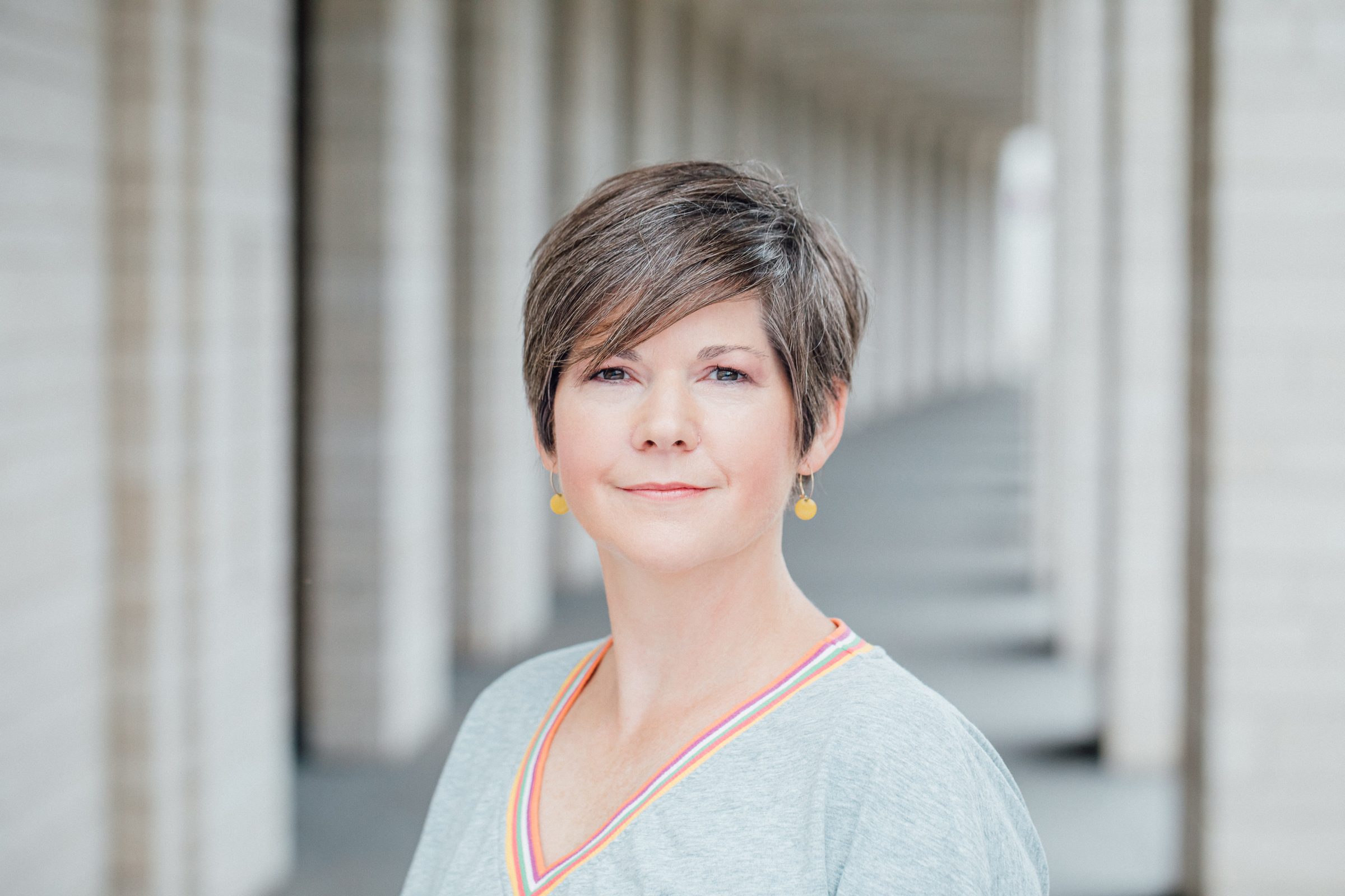 Porträt einer Frau mit kurzen braunen Haaren und gelben Ohrringen in einem hellen Säulengang, Fotoshooting für Colli Natur Friseure