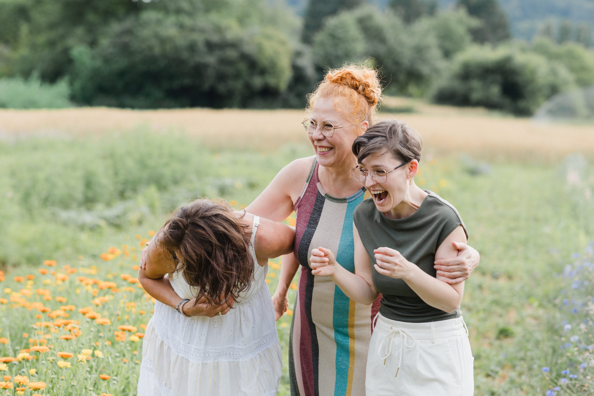 Drei Frauen lachen ausgelassen auf einer Wiese mit orangefarbenen Blüten, eine beugt sich vor Lachen nach vorn, Team von Colli Natur Friseure