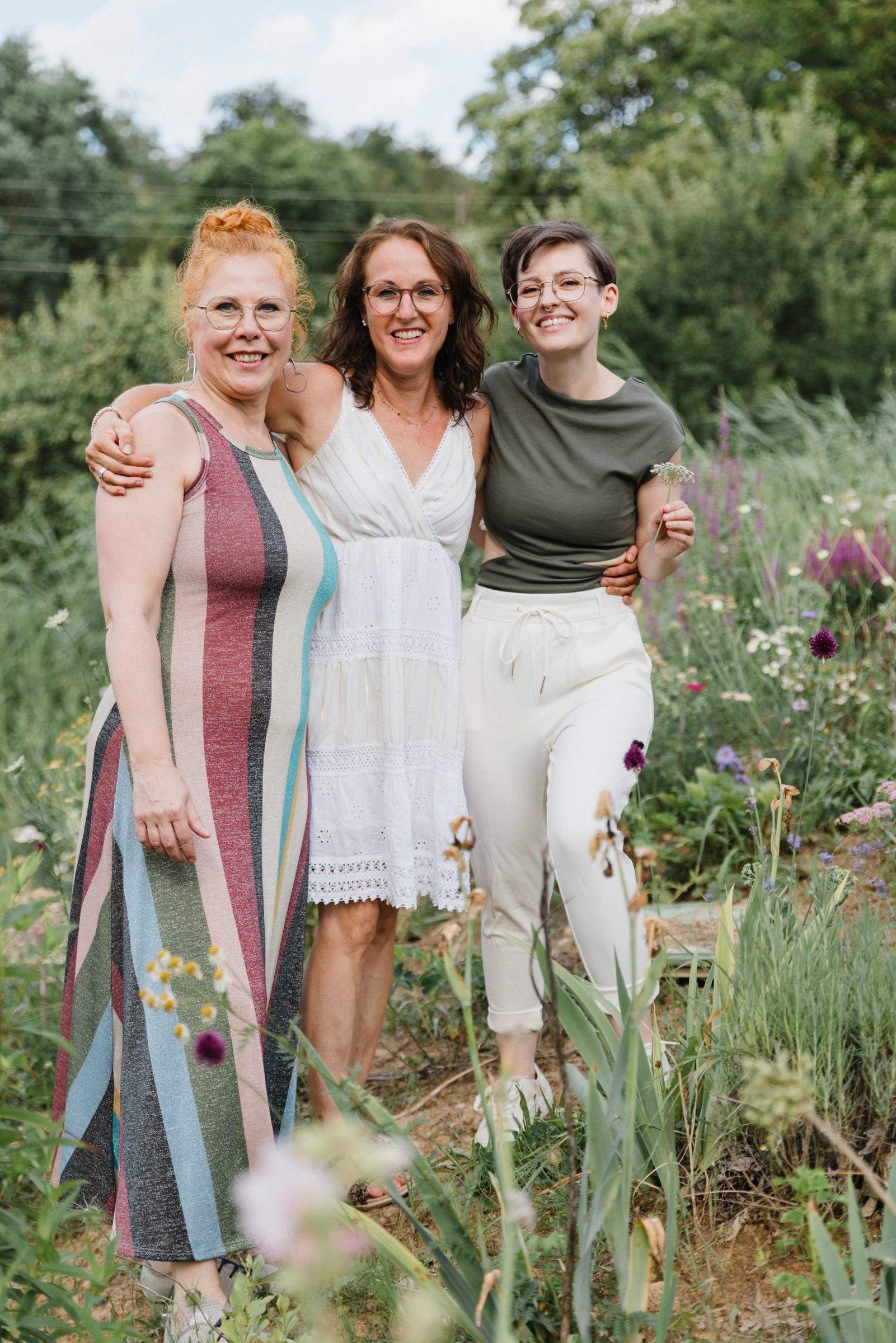 Drei Frauen stehen Arm in Arm lächelnd in einem blühenden Garten, Team von Colli Natur Friseure
