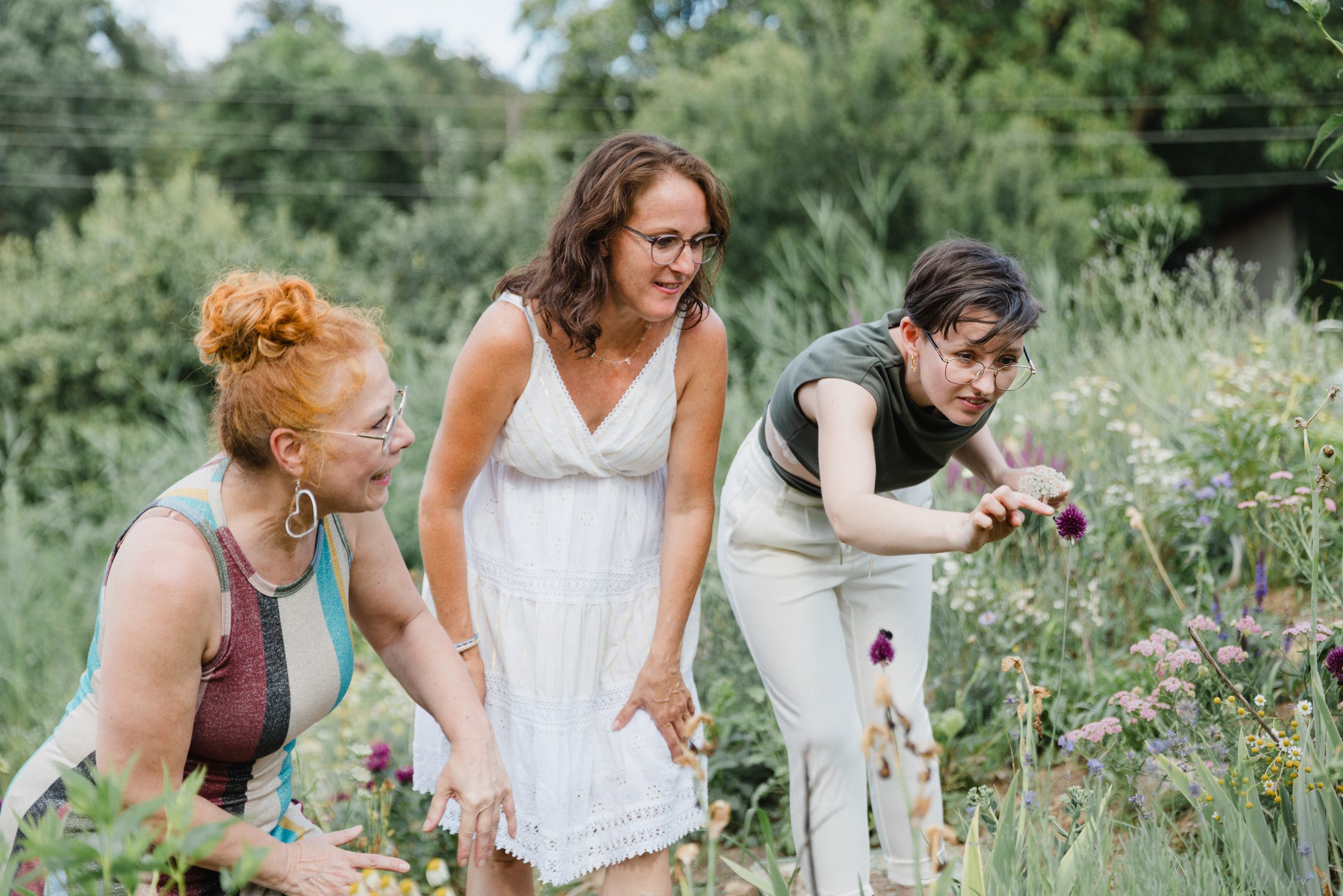 Drei Frauen beugen sich lächelnd über Wildblumen in einem Garten und betrachten die Blüten, Team von Colli Natur Friseure