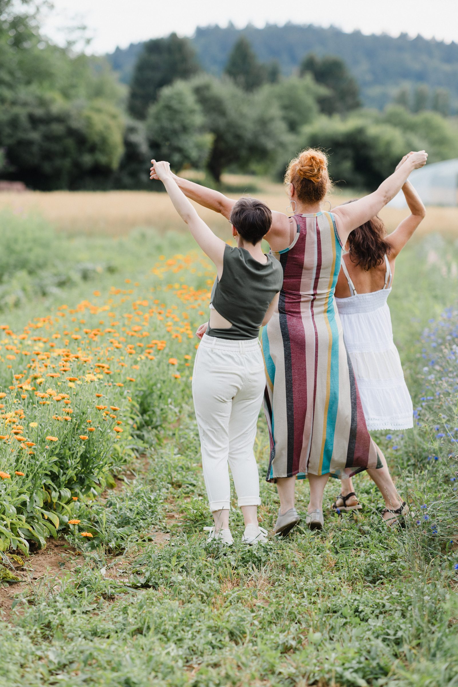 Drei Frauen strecken auf einer Wiese mit orangefarbenen Blüten fröhlich die Arme in die Luft, Rückenansicht, Team von Colli Natur Friseure