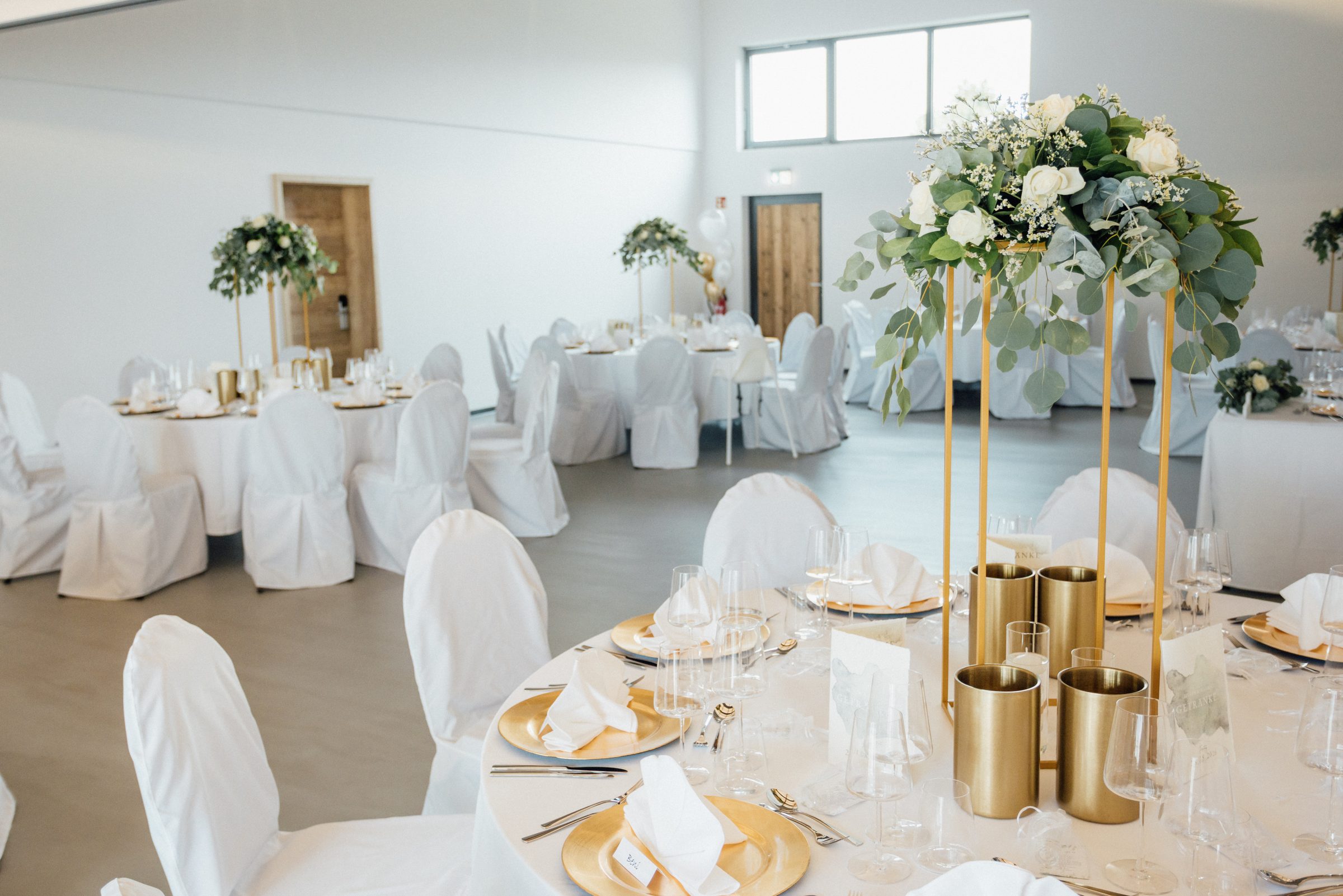 Zur Hochzeit festlich eingedeckter Saal im Eventum des Hotel Engelhorn mit weißen Stuhlhussen, goldenen Platztellern und Blumengestecken