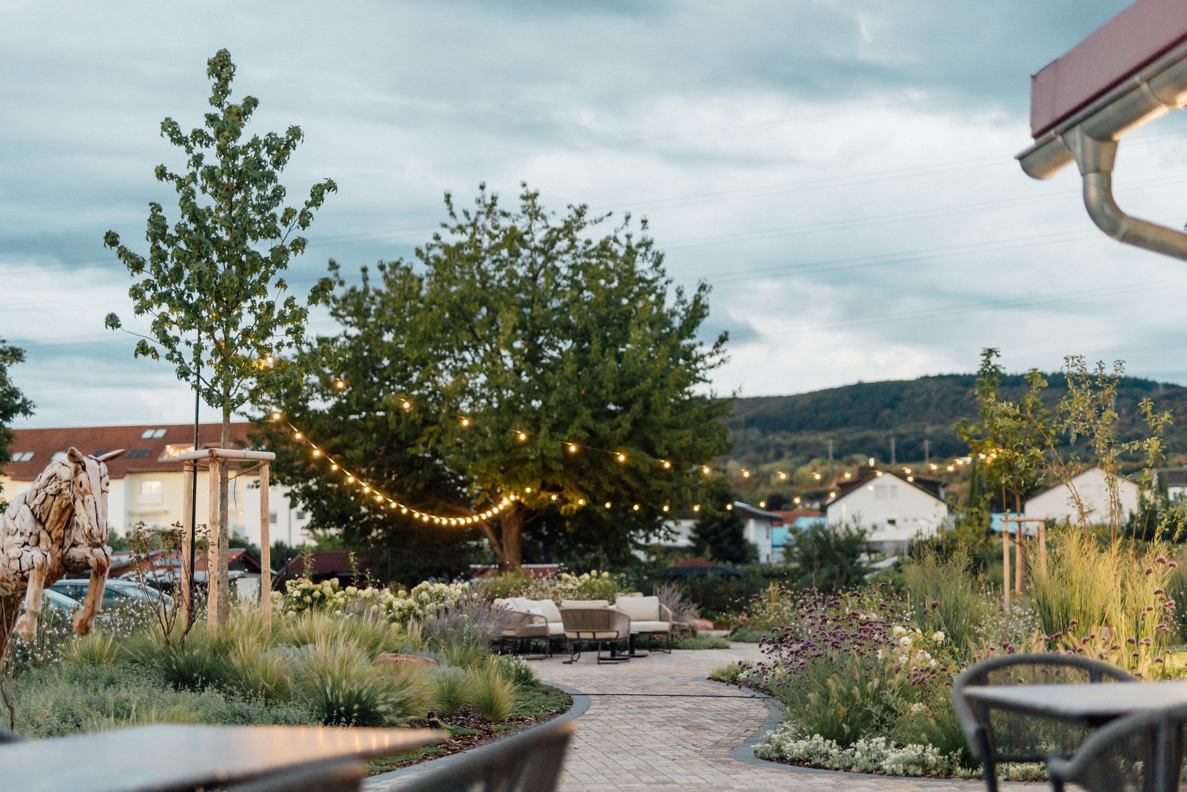Garten des Eventum beim Hotel Engelhorn in der Abenddämmerung mit Lichterketten, Holzpferd, Gräsern und Lounge-Möbeln