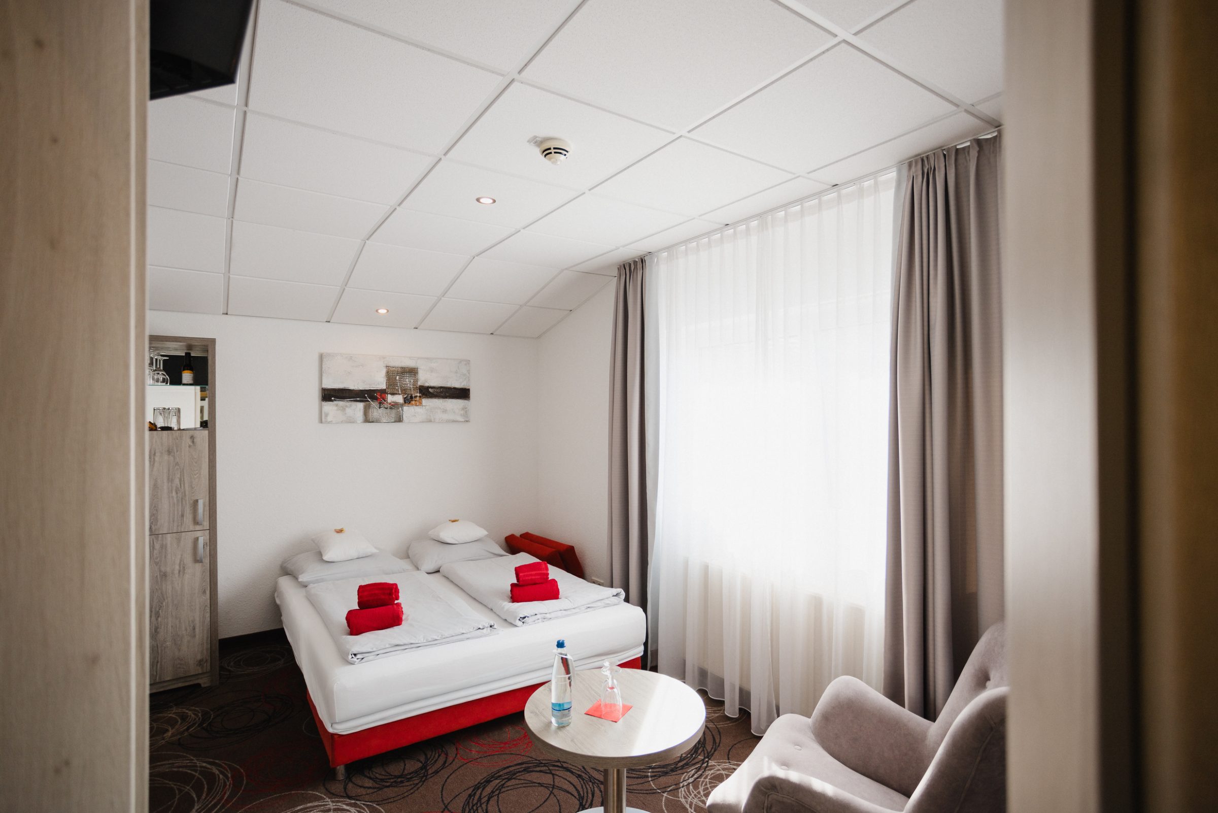 Doppelzimmer im Hotel Engelhorn mit roten Handtüchern auf dem Bett, Sessel, Beistelltisch und hellen Vorhängen