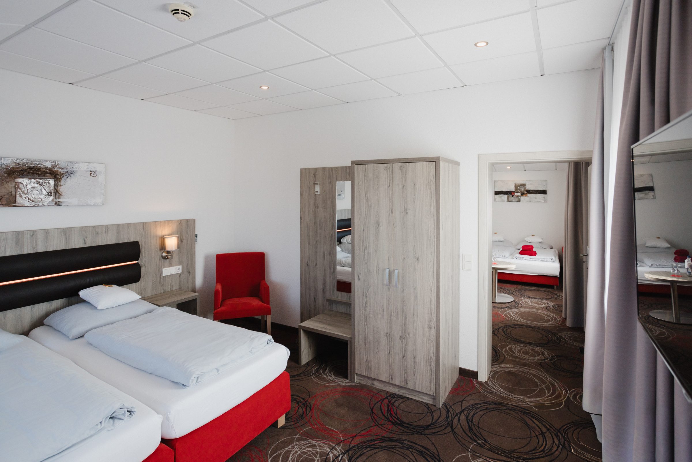 Doppelzimmer im Hotel Engelhorn mit gepolstertem Kopfteil, rotem Sessel und Kleiderschrank, im Hintergrund ein weiteres Zimmer