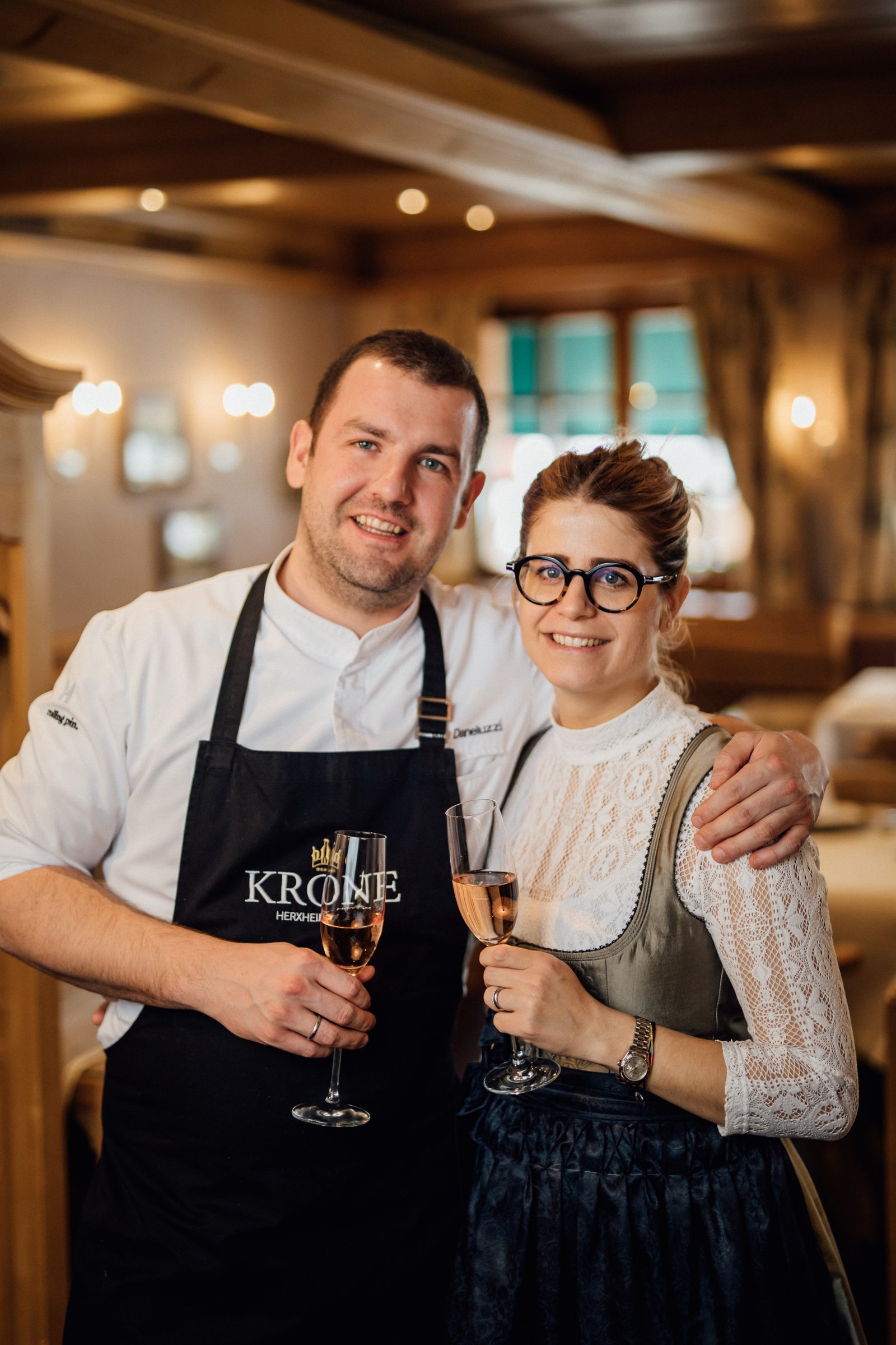 Koch und Gastgeberin im Dirndl stehen lächelnd mit Sektgläsern im Restaurant des Hotel Krone