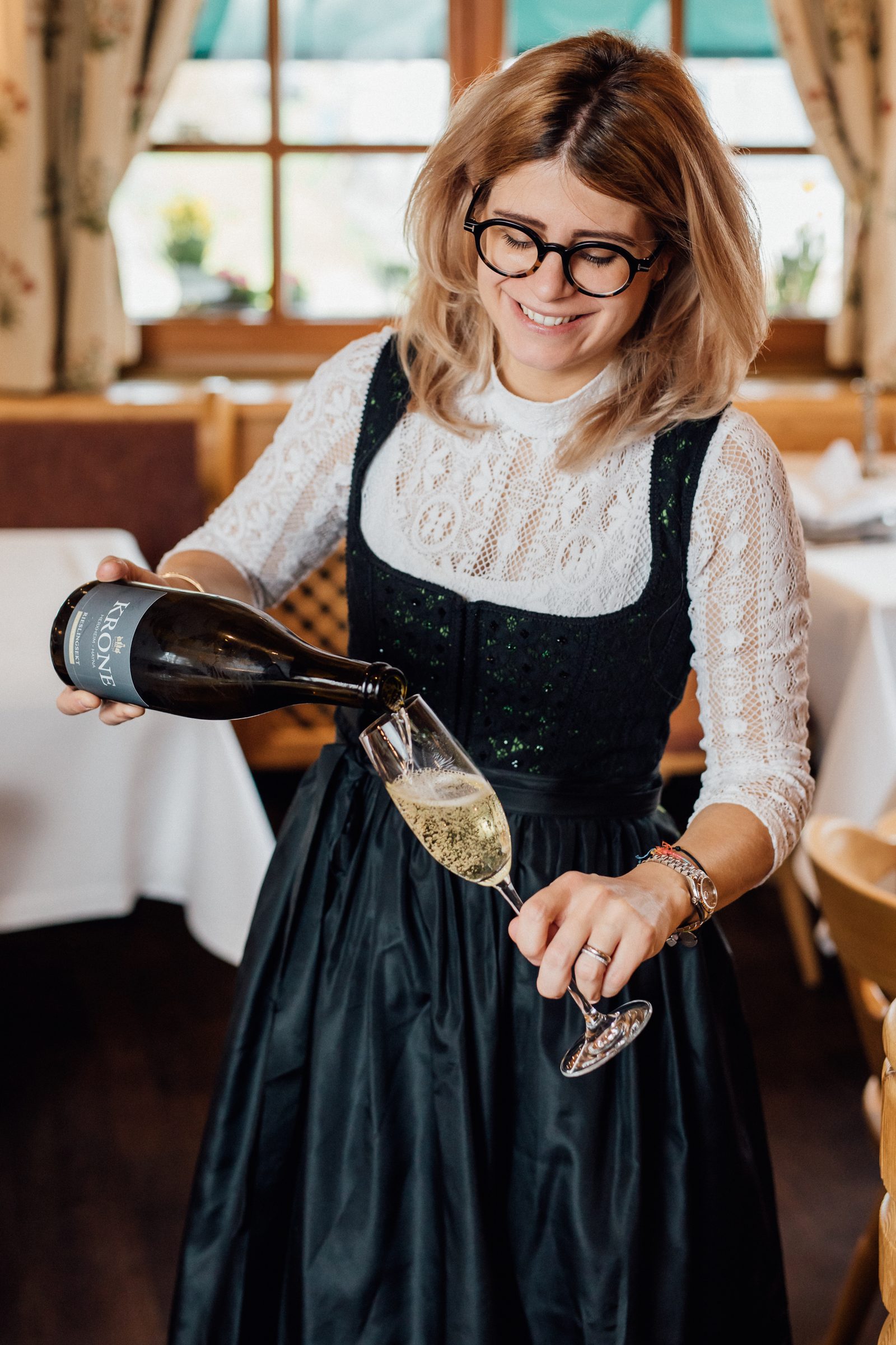 Gastgeberin im Dirndl schenkt im Restaurant des Hotel Krone lächelnd Sekt in ein Glas ein
