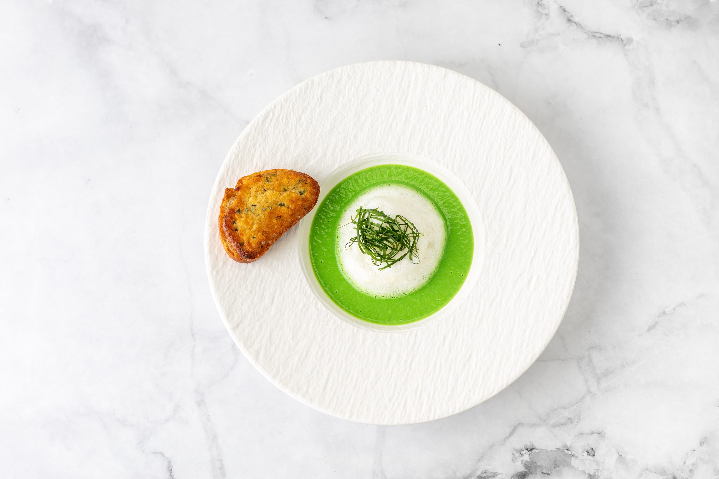 Helle Suppe mit grünem Rand und Schnittlauch, daneben ein geröstetes Brot, auf einem weißen Teller auf Marmor, von oben fotografiert