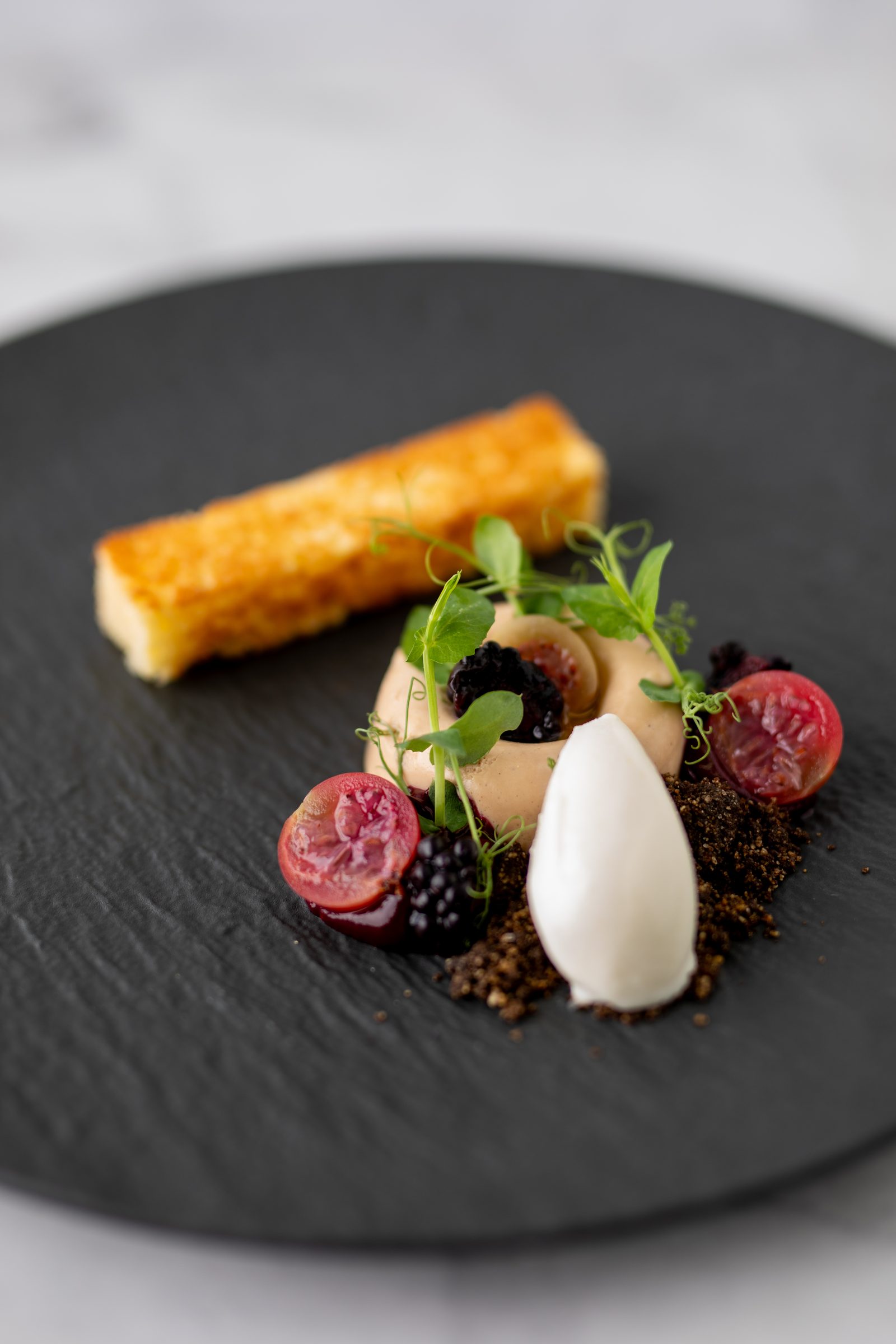 Dessert mit Eisnocke, Beeren, Schokoladenkrümeln, Kräutern und einem knusprigen Riegel auf einem schwarzen Teller