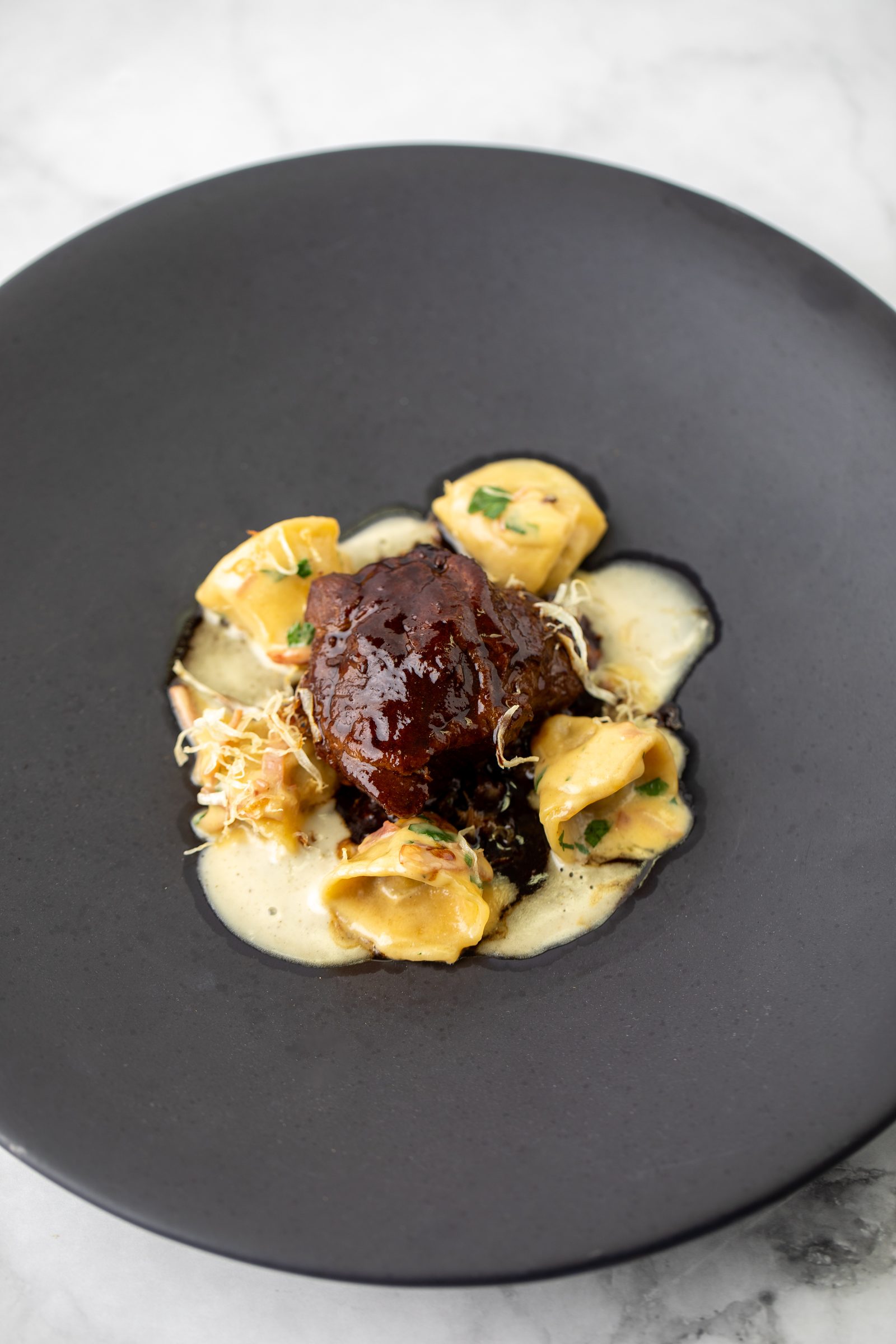 Geschmortes Fleisch mit Tortellini, heller Sauce und frittierten Zwiebeln auf einem dunklen Teller