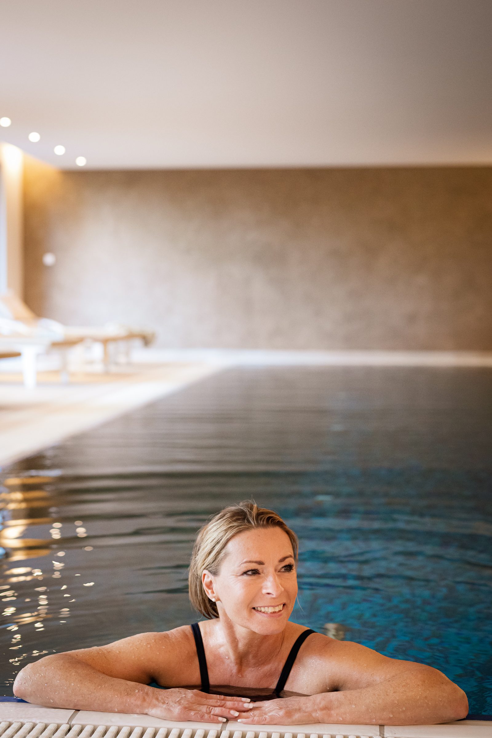 Frau im Badeanzug stützt sich lächelnd auf den Rand des Innenpools im Hotel Krone, dahinter Liegen und eine Natursteinwand