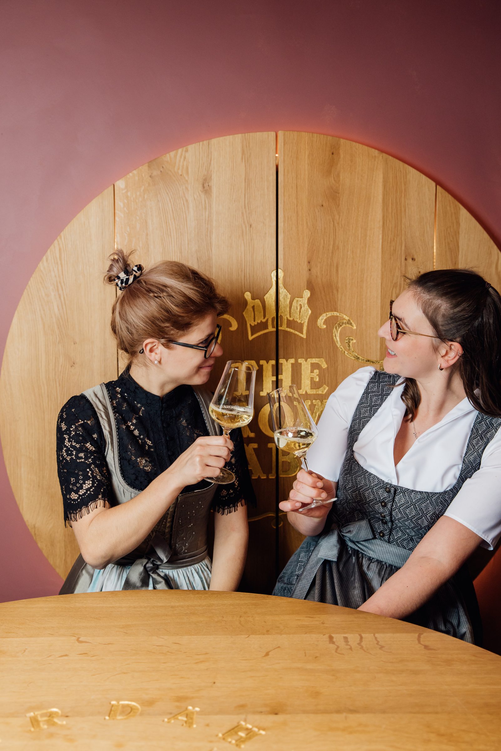 Zwei Mitarbeiterinnen im Dirndl stoßen mit Weißwein an einem runden Holztisch an, dahinter eine Holzwand mit goldener Krone