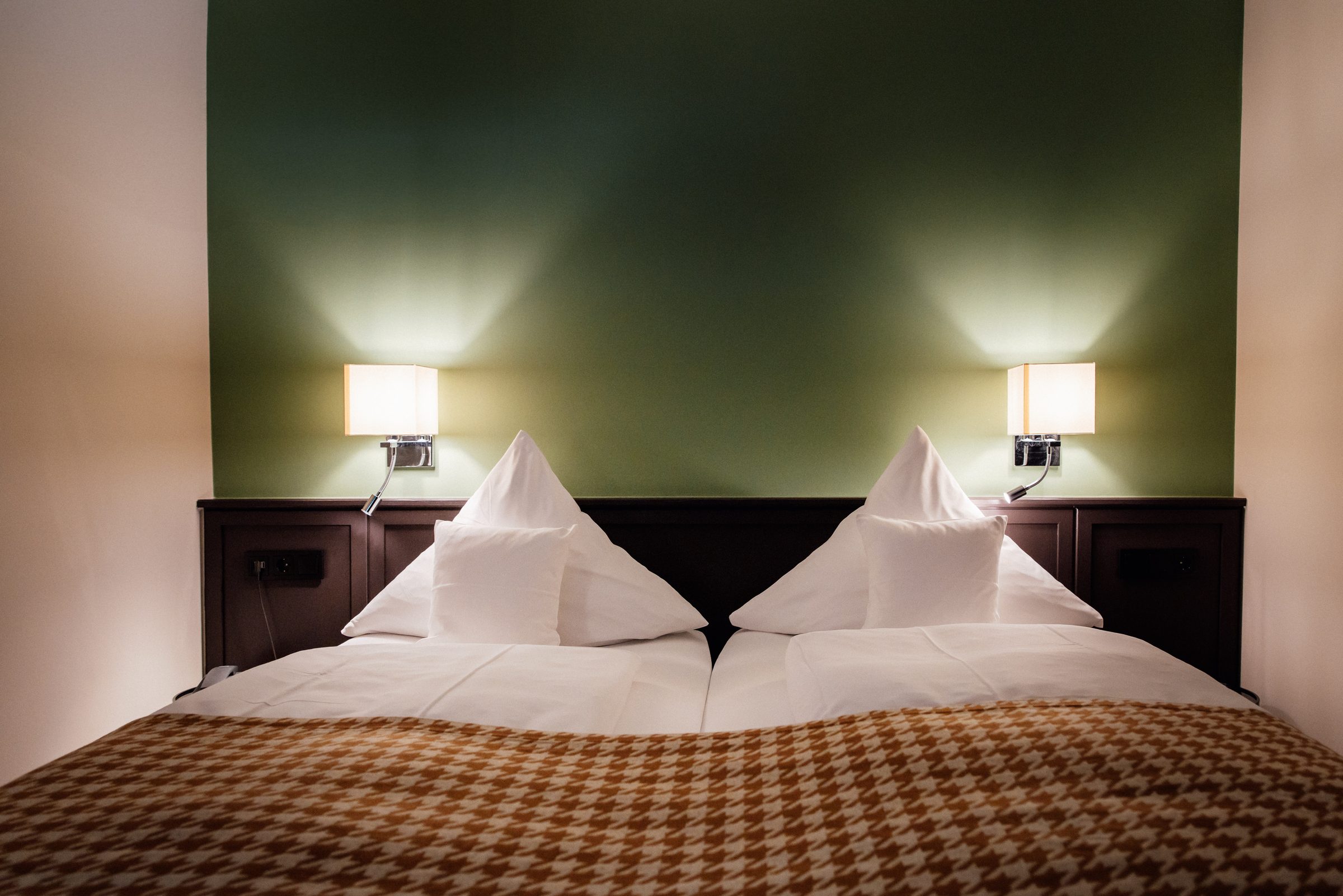 Renoviertes Doppelzimmer im Hotel Krone mit dunkelgrüner Wand, zwei Leselampen und einer Tagesdecke im Hahnentrittmuster