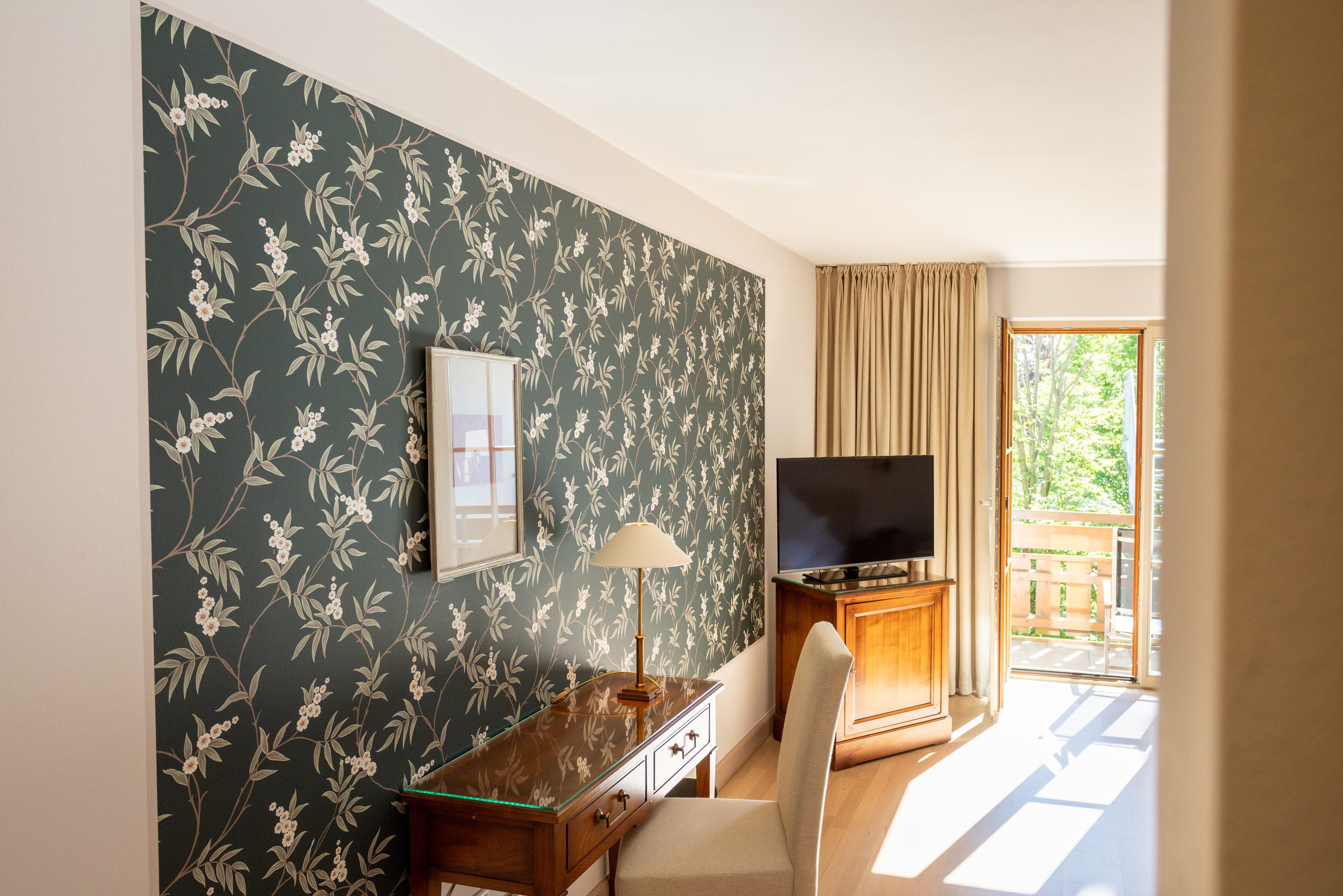 Hotelzimmer im Hotel Krone mit dunkler Blumentapete, Schreibtisch, Fernseher und offener Balkontür ins Grüne