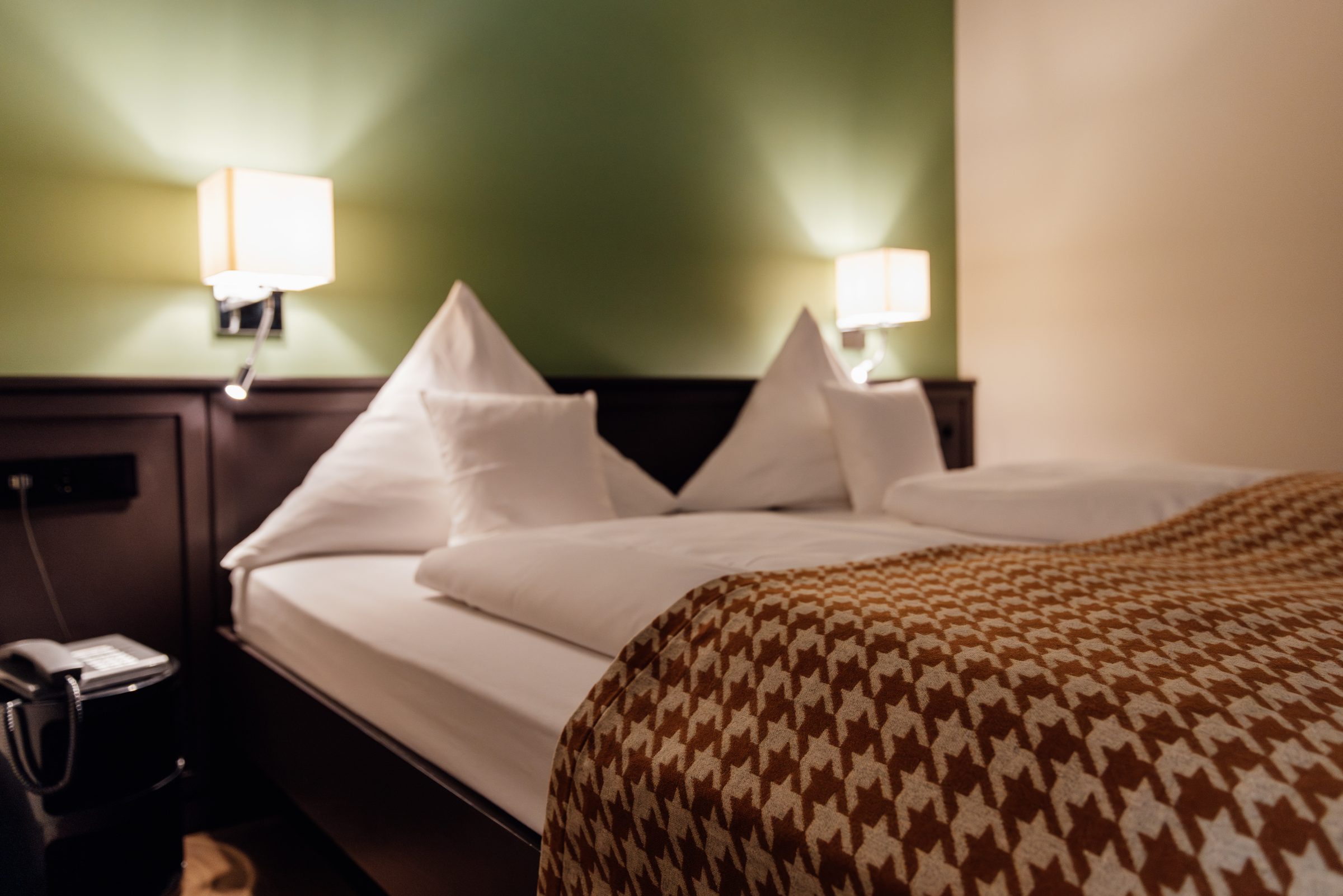 Doppelbett mit Tagesdecke im Hahnentrittmuster und zwei Leselampen vor einer dunkelgrünen Wand im Hotel Krone