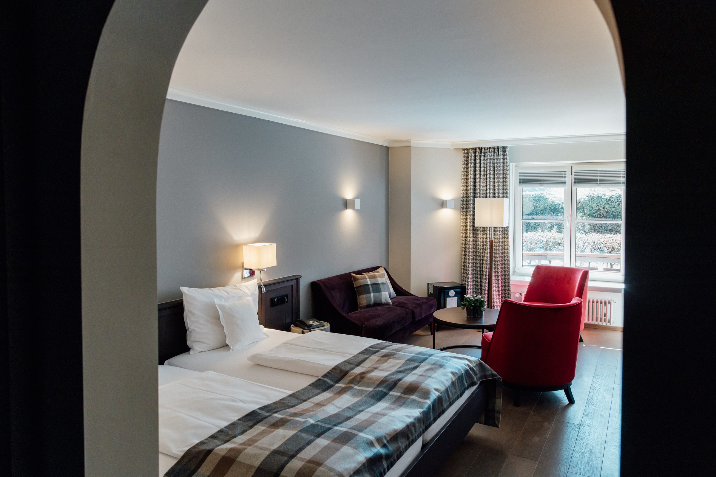 Blick durch einen Rundbogen in ein Doppelzimmer im Hotel Krone mit karierter Tagesdecke, rotem Sessel und Sofa am Fenster
