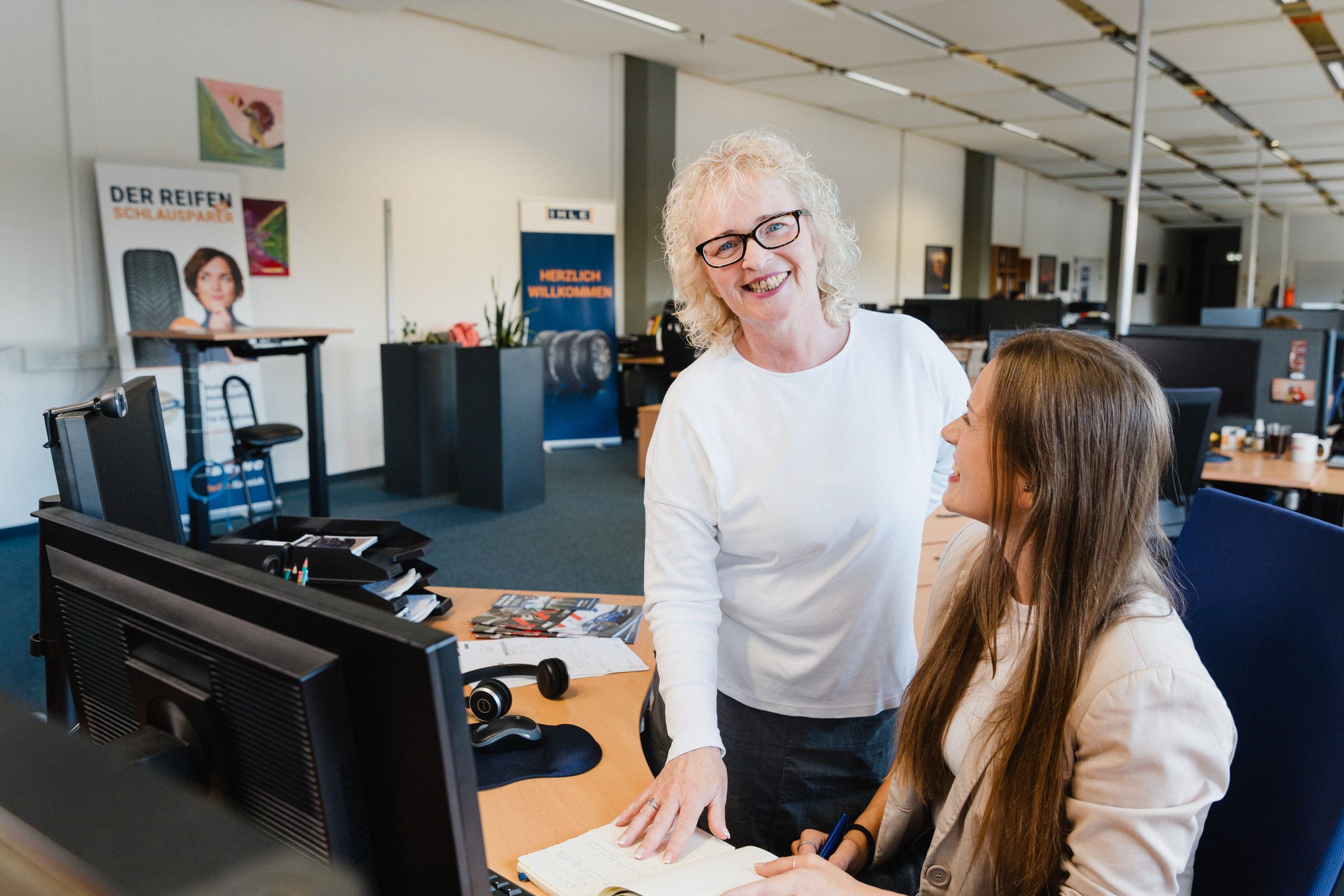 Zwei Mitarbeiterinnen von IHLE unterhalten sich lachend an einem Arbeitsplatz im Großraumbüro