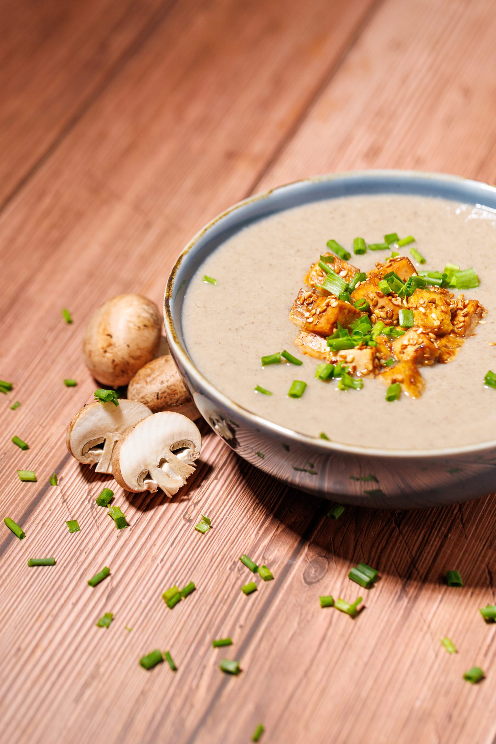 Champignon-Cremesuppe mit gebratenem Tofu und Schnittlauch in einer Schale auf einem Holztisch, daneben frische Champignons, Rezeptfoto für ein GU-Kochbuch