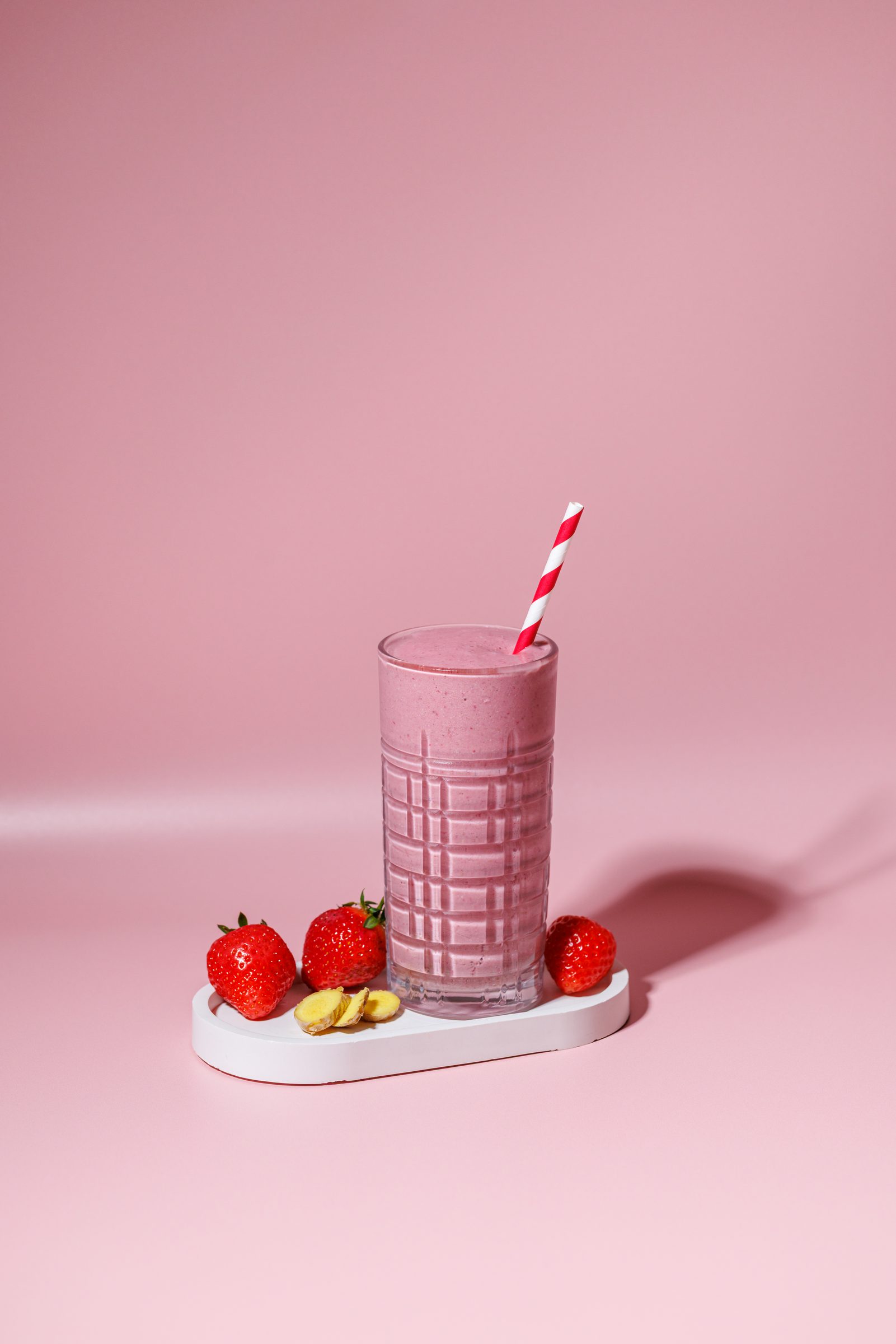 Rosa Smoothie im Glas mit rot-weißem Strohhalm vor rosa Hintergrund, davor frische Erdbeeren, Rezeptfoto für ein GU-Kochbuch