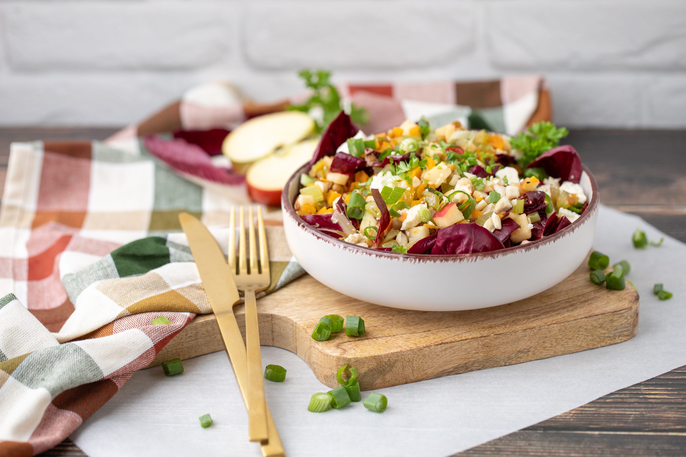 Linsen-Apfel-Salat mit Radicchio und Feta in einer Schale auf einem Holzbrett, daneben goldenes Besteck und ein karierter Stoff