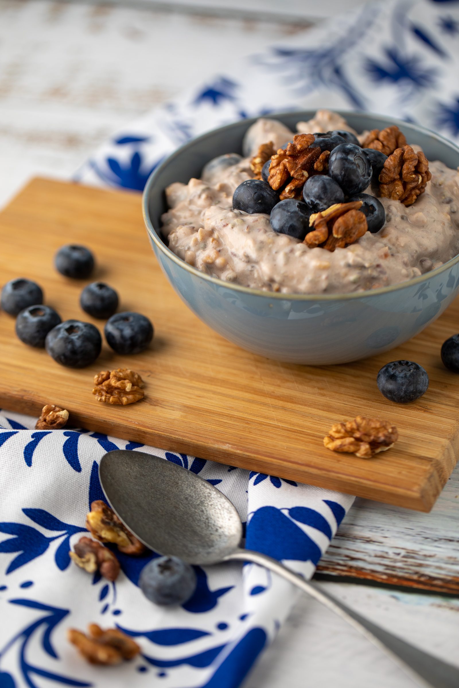 Schale mit Porridge, Heidelbeeren und Walnüssen auf einem Holzbrett und einem blau gemusterten Tuch