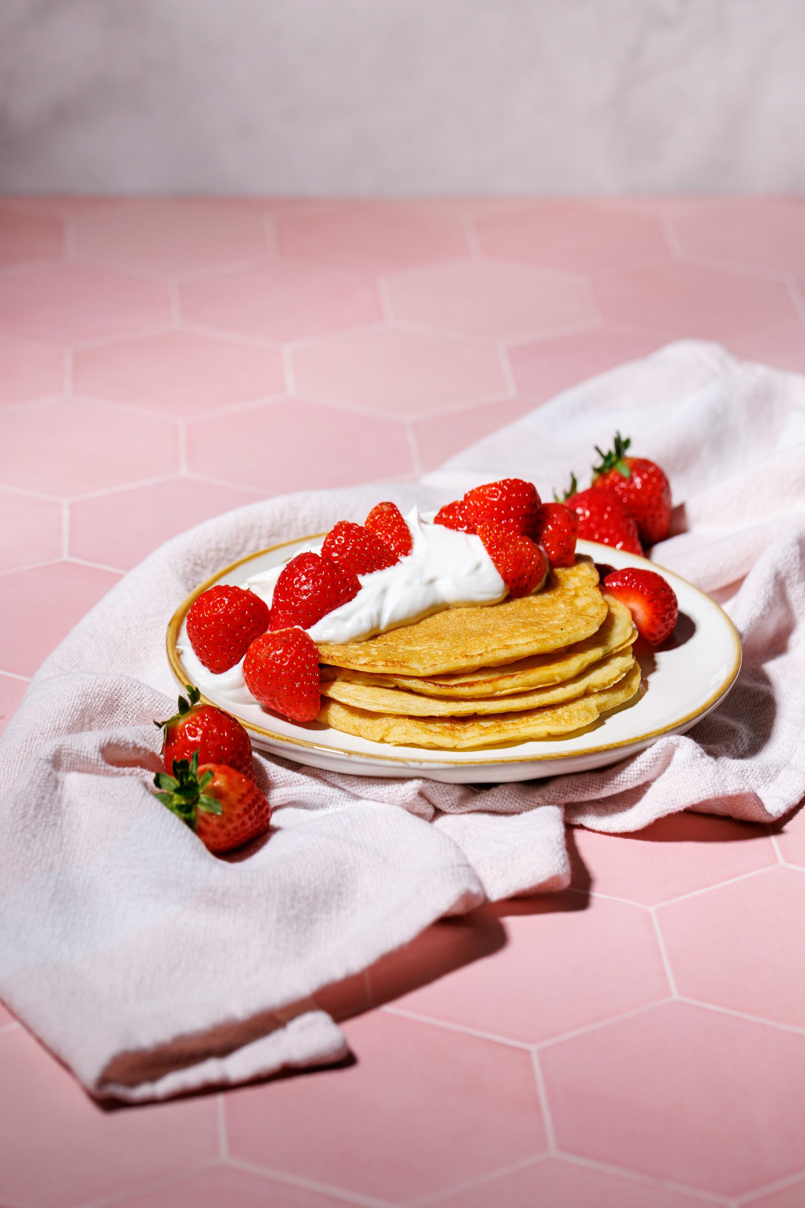 Pancakes mit Joghurt und Erdbeeren auf einem Teller vor rosa Fliesen, Rezeptfoto für ein GU-Kochbuch
