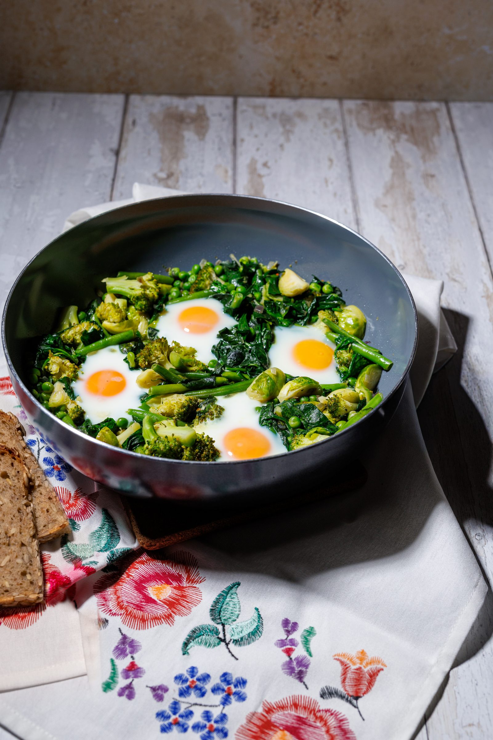 Grünes Shakshuka mit Spiegeleiern und Brokkoli in einer Pfanne auf einem bestickten Tuch, daneben Brotscheiben, Rezeptfoto für ein GU-Kochbuch