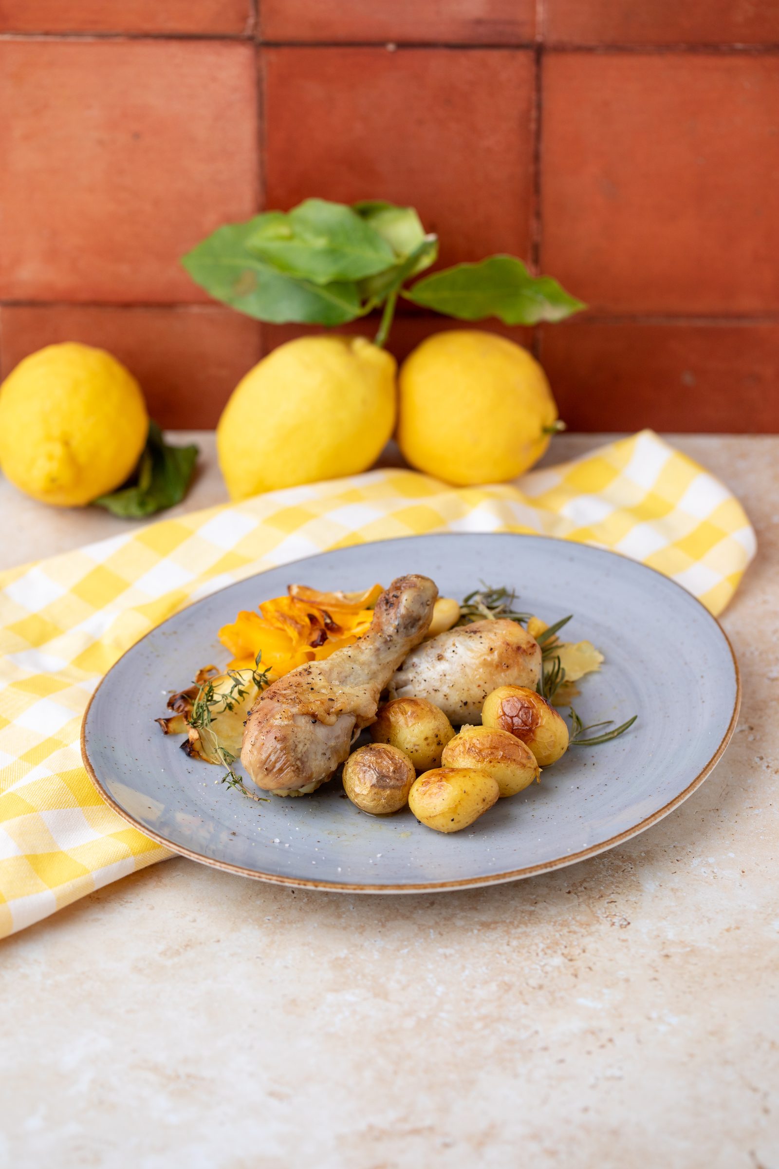 Hähnchenkeulen mit Kräutern und kleinen Kartoffeln auf einem grauen Teller, dahinter Zitronen vor Terrakottafliesen, Rezeptfoto für ein GU-Kochbuch