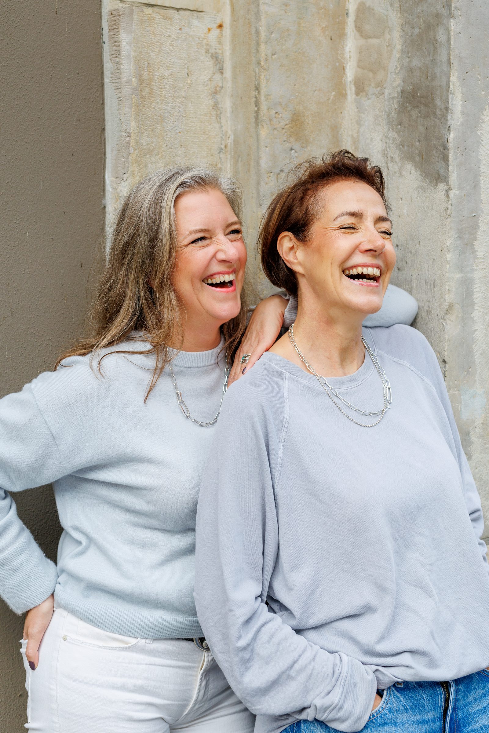 Zwei Frauen mit Silberketten von Lilienglück lachen gemeinsam an einer Steinwand