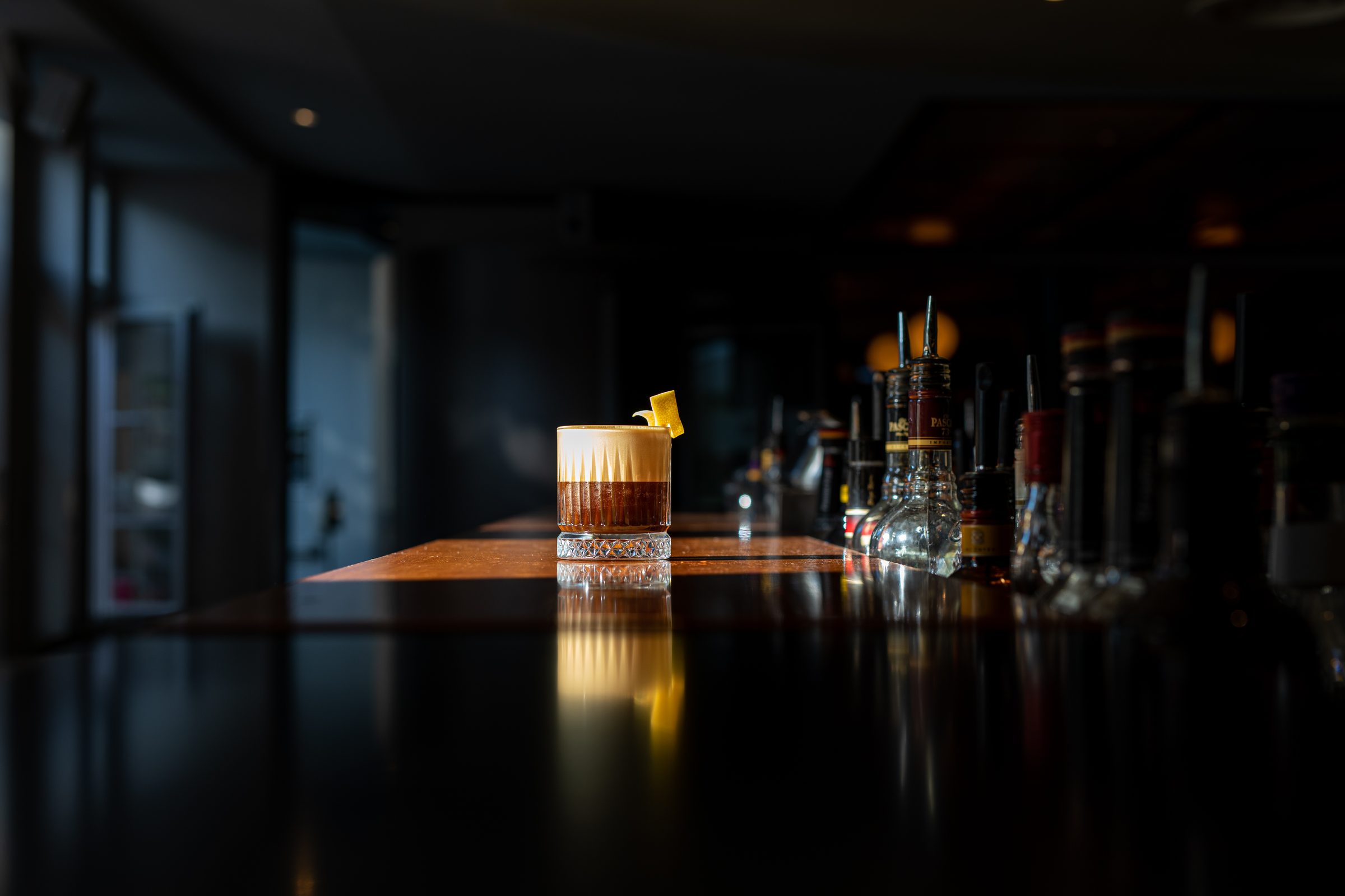 Geschichteter Drink mit heller Schaumschicht und Zitronenzeste im Tumbler auf dem spiegelnden Tresen der dunklen Bar des Ludwig in Mosbach