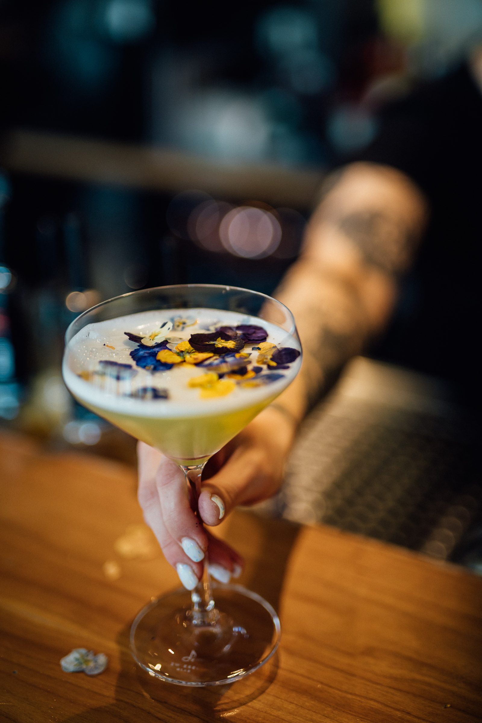 Hand reicht einen hellen Cocktail mit essbaren Blüten über den Holztresen der Bar des Ludwig in Mosbach